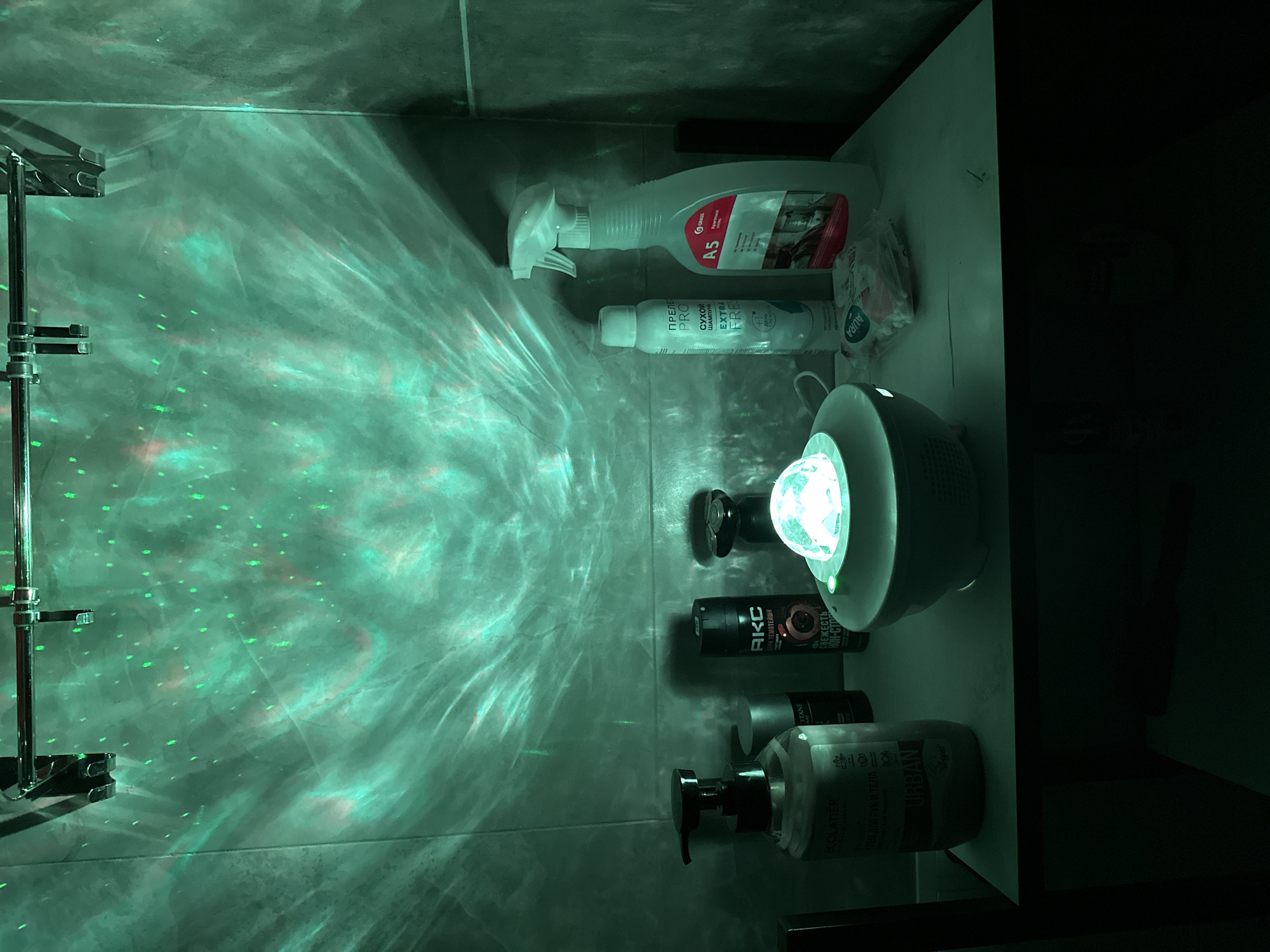 The scene is a brightly lit bathroom, possibly in Sochi, Russia, given the Russian language on the product labels. The walls are tiled in a dark grey color. On a white countertop are several personal care and cleaning products: a spray bottle labeled "A5", a dry shampoo bottle, a hand and body lotion labeled "URBAN", a bottle of axe body spray and a lamp or projector that is the source of the bright light and scattered green patterns on the walls. There's also a metal shower rack or fixture visible on the left side of the frame. The right side of the image fades into darkness, suggesting it may be a corner of the bathroom.