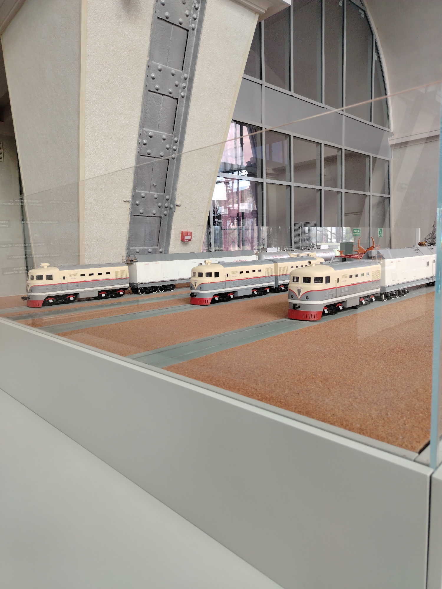 This media file shows a detailed model display of trains in what appears to be a museum or exhibition setting in Moscow, Russia. The scene features three model trains on a track, with a backdrop of a large building interior that includes exposed structural elements like beams and rivets, along with large glass windows. The trains are designed to resemble older diesel locomotives, with a cream, grey, and red color scheme. The first train in the foreground has a distinctive rounded front and windows along its side. Behind it, another train consists of a locomotive and a white cargo car. Further back, another similar train is visible. The track area is depicted with a textured, light brown surface. The overall lighting suggests an indoor environment, possibly daytime given the light coming through the windows, though no specific time of day can be determined. The setting implies an exhibit about transportation or industrial history. There is no visible text in the main view that is legible.