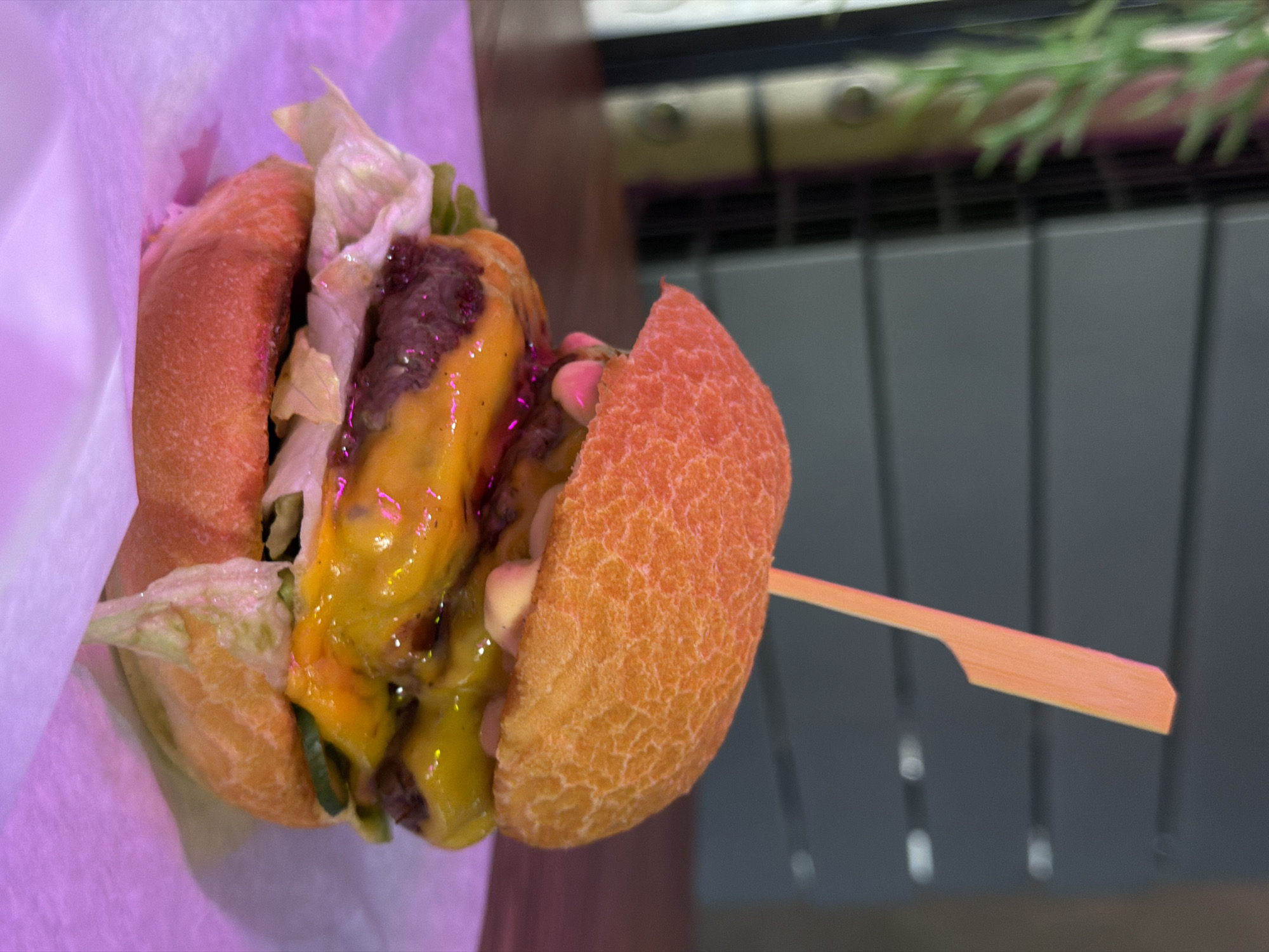 A close-up shot depicts a hamburger held together by a wooden skewer. The hamburger consists of a bun, lettuce, what appears to be a meat patty, and melted cheese. It is partially wrapped in white paper. The background features what appears to be a blurred metal structure with green vegetation visible at the top. The setting is indoors and lit with a faint purple hue. The location and city cannot be determined.