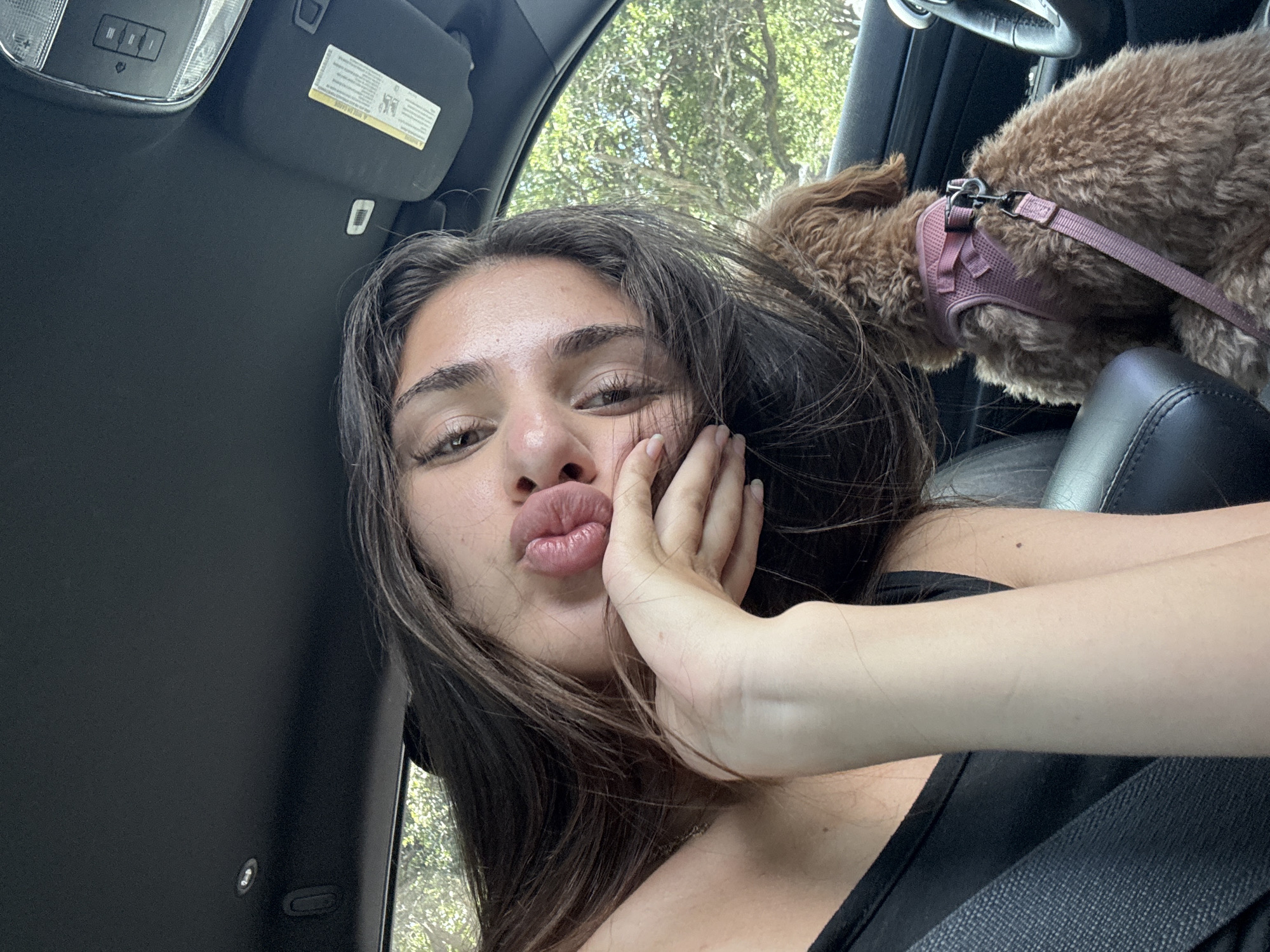 A woman is in the driver's seat of a car, with a dog on the passenger seat. The woman has dark hair and is wearing a black tank top. She is making a kissing face and has one hand on her cheek. The dog is brown and wearing a pink harness. The car is parked in front of trees, in the city of Delray Beach, United States of America.