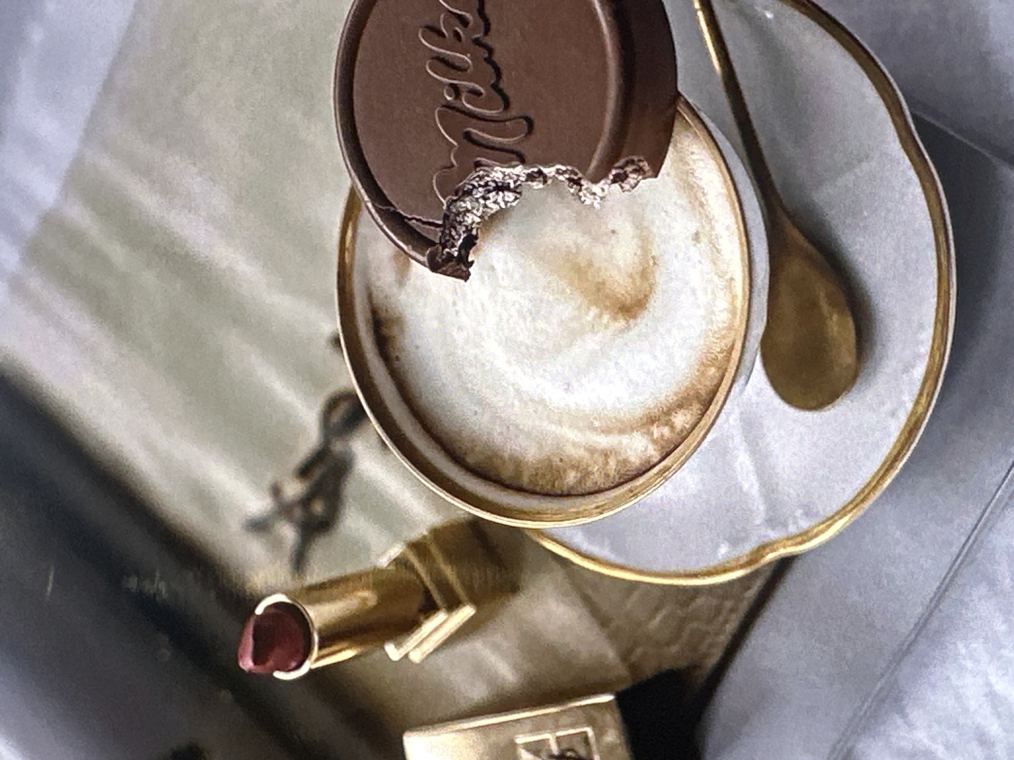 A close-up overhead shot shows a cup of latte with a bitten Milka chocolate coin resting on the rim. A gold spoon lies beside the cup on a saucer with a gold trim. A gold YSL lipstick sits near the bottom of the image. The setting suggests an indoor café or home environment. The location in Yerevan, Armenia, is not definitively inferable from the image alone.