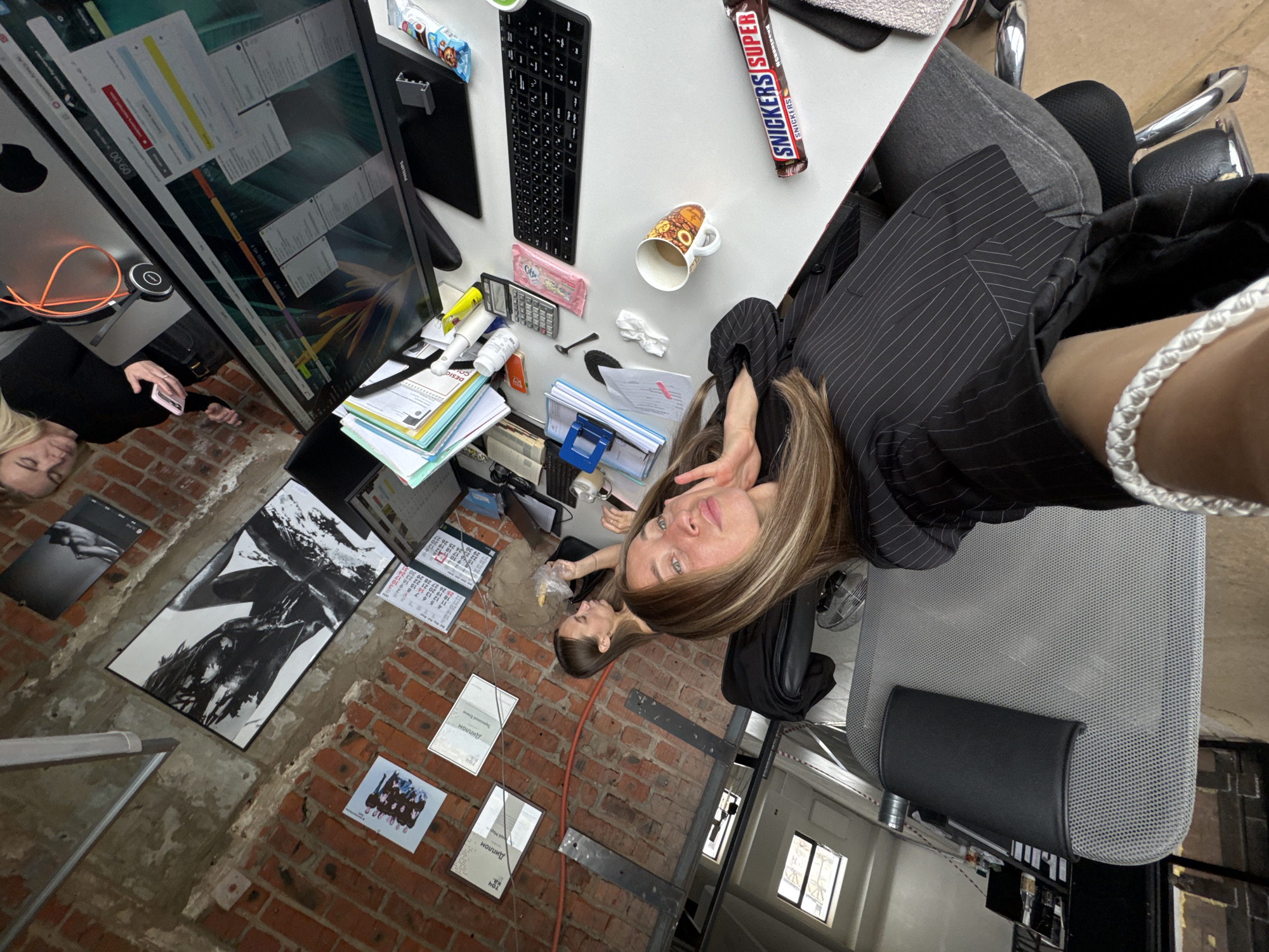 This media file captures an office environment from a high, rotated perspective, showcasing two women and a busy workspace. The main subject, a woman with long, highlighted hair and a dark pinstriped jacket, looks directly at the camera with a neutral expression, revealing a white braided bracelet on her wrist. Another woman with blonde hair is visible to the left, appearing to interact with a phone.
The white desk is cluttered with a Philips computer monitor displaying "09:00" and numerical data like "28 632 145 P". Nearby are a black keyboard, Snickers Super and Snickers chocolate bars, a patterned ceramic mug, a calculator, a pink packet, a crumpled tissue, a yellow marker, a book marked "DESIC", and various stacks of papers and office supplies. A grey mesh office chair is also present.
The floor, characterized by exposed red brick and concrete, holds several items: two documents labeled "Диплом" (Diploma), a large black and white framed artwork featuring a person, smaller prints, and calendar pages displaying numbers such as "18 19 2" and "25 26 29". An orange cable trails across the floor. The setting suggests an active, informal workspace, likely within an older building. The visible Russian text confirms the location context of Chaikovsky, Russia.