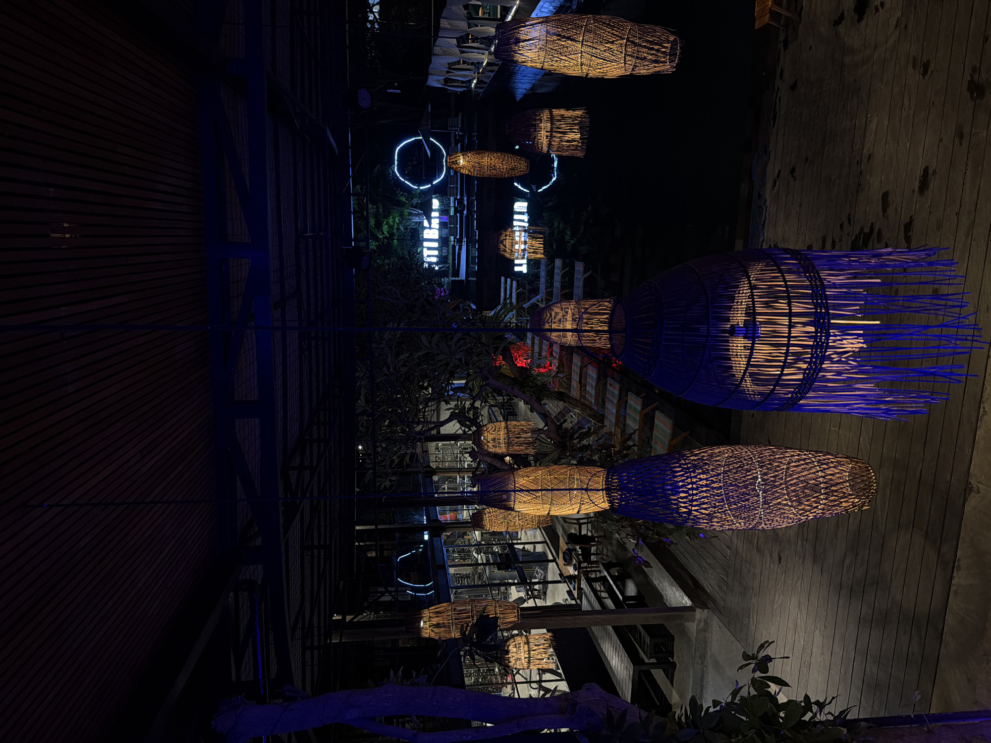 This image captures an outdoor setting at night in Ubud District, Indonesia, featuring decorative, woven lampshades hanging from above. The lighting scheme casts a blue and yellow glow on the scene, emphasizing the texture of the lampshades and the surrounding foliage. Reflections of the lampshades and neon lights are visible in a nearby body of water. A wooden structure and deck are present in the foreground and mid-ground, indicating an outdoor dining or relaxation area. Some plants are visible at the bottom of the frame, and the picture is rotated to its side for some reason.