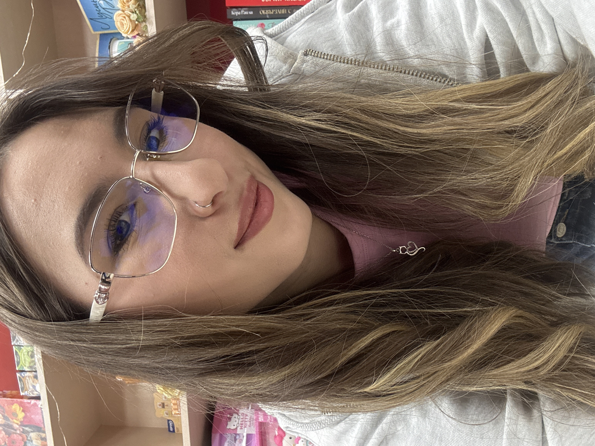 A close-up portrait, taken at a slight tilt, features a young woman with long, layered light brown hair streaked with blonde highlights. She looks directly at the viewer with a calm expression, her lips slightly parted in what appears to be a gentle smile. She is wearing silver-rimmed glasses that reflect blue light, possibly indicating anti-blue light lenses, and has a small silver hoop nose piercing on her right nostril. Her eyes are blue.
She is dressed in a light pink top, partially visible beneath a light grey zippered hoodie. A delicate silver necklace with a heart-shaped pendant rests on her collarbone. The background suggests an indoor setting, likely a shop or a room with shelves. Behind her, various objects are visible, including stacked books. Partially legible text on a blue decorative item reads "Любов" (Lyubov, meaning "Love" in Bulgarian), and book titles include "Първа Рана" (Parva Rana, "First Wound") and "ВЪОБРАЗИМ С" (Vaobrazim S, "Imaginary S"). Other smaller items or products are also present on shelves in the lower background. The scene appears brightly lit. This image is contextualized as being from Veliko Tarnovo, Bulgaria.