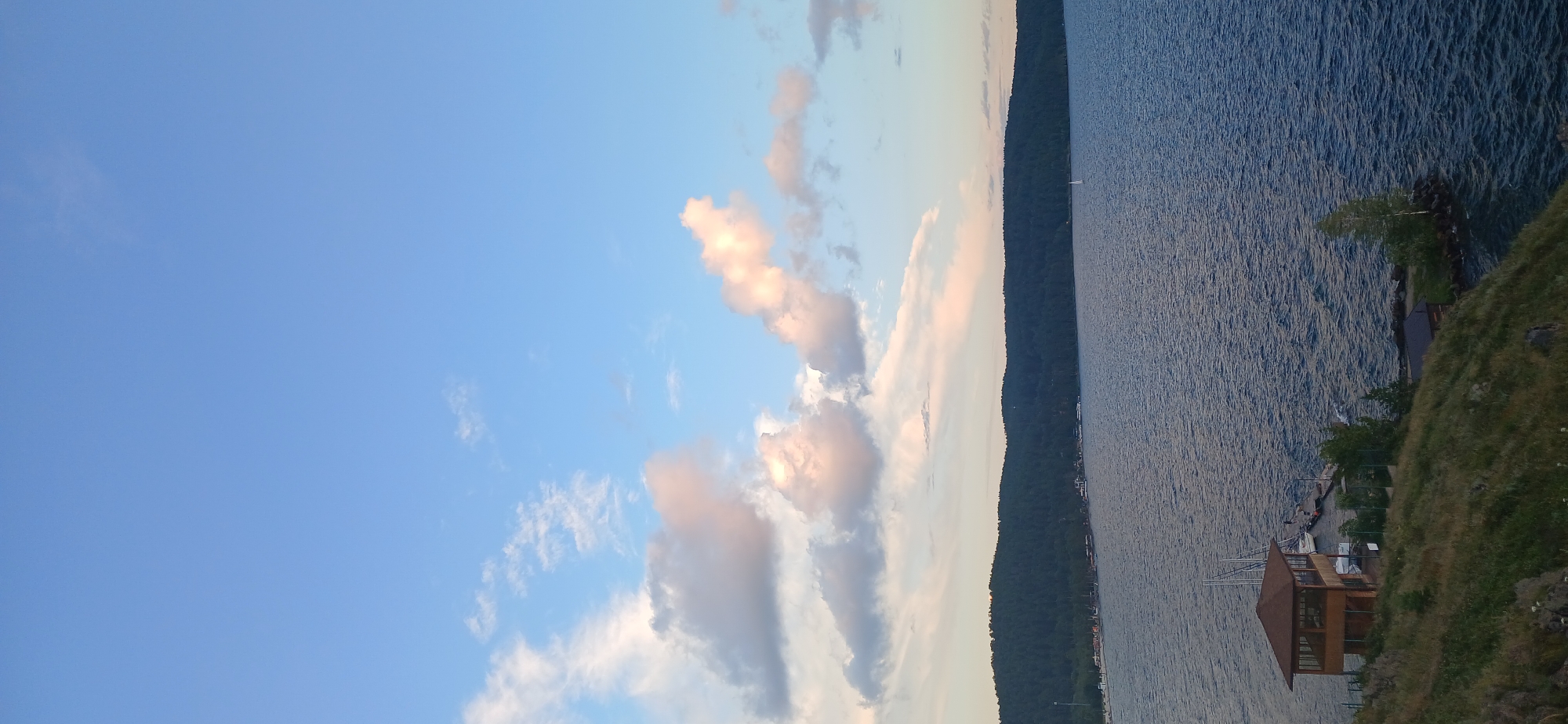 This image captures a scenic view of Lake Turgoyak in Turgoyak, Russia, during what appears to be late afternoon or early evening. The sky is a vibrant blue, dotted with scattered clouds that are tinged with soft pink and orange hues, suggesting the approach of sunset.
The foreground features a grassy hillside, with some patches of dry grass and small rocks. On this hillside, a small, wooden boathouse with a brown roof and glass windows is situated. A dock extends from the shore to the boathouse, where several sailboats are moored. Further down the hill, closer to the water's edge, another small structure with a dark roof is visible.
The middle ground is dominated by the expansive, blue waters of Lake Turgoyak. The surface of the lake is rippled, indicating a gentle breeze. In the distance, a long, densely forested hill forms the horizon, creating a dark, verdant backdrop against the sky. Along the shoreline of this hill, there are signs of civilization, with small buildings and what appears to be a beach or recreational area. A lone white sailboat is visible on the lake in the distance. The overall atmosphere is serene and picturesque.