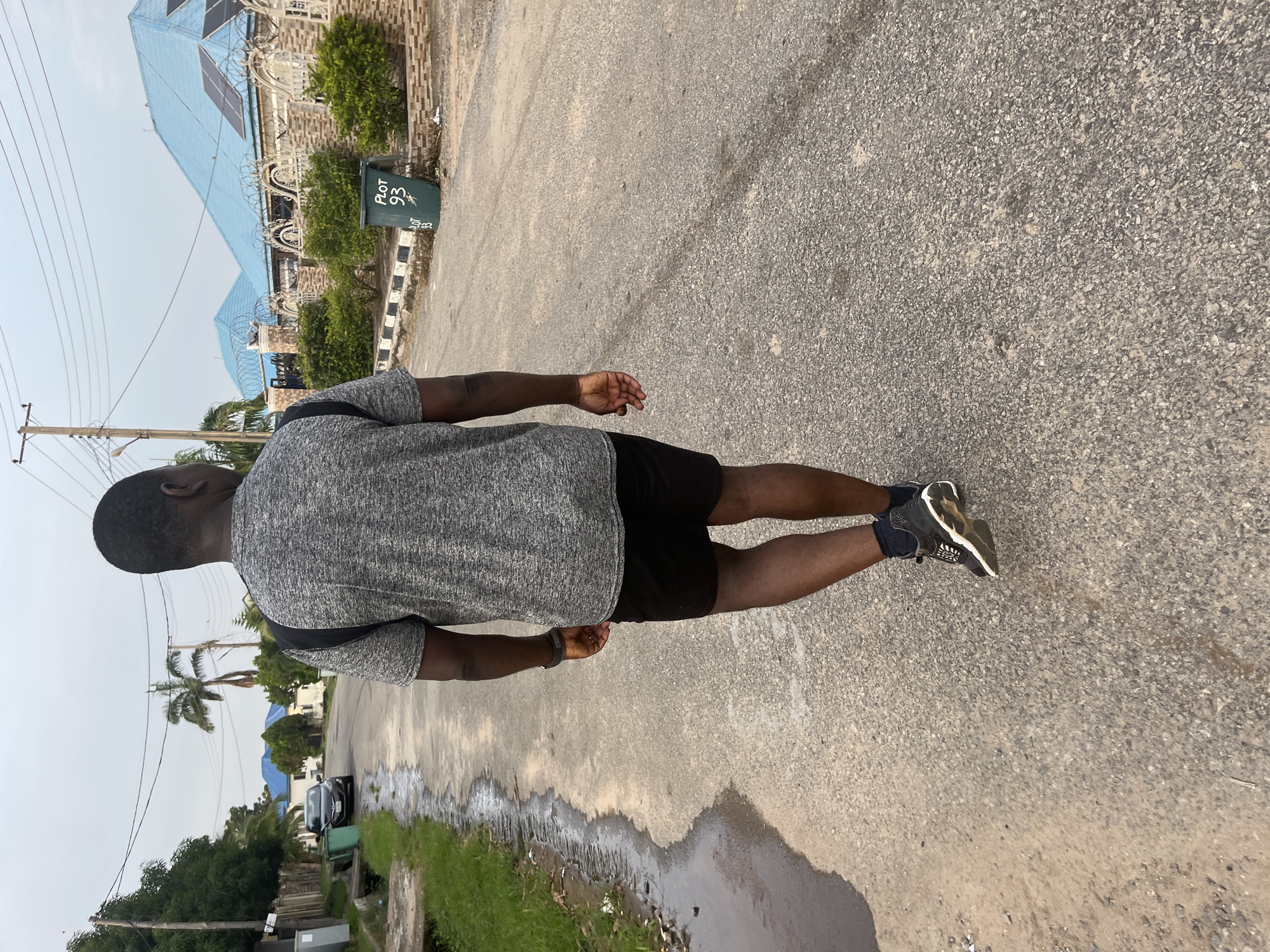 A man walks away from the camera on a paved road in Abuja, Nigeria. The man is wearing a gray t-shirt with black trim, black shorts, and black and white athletic shoes. He has a black backpack on his shoulders. The road is wet in places, indicating recent rain. To the left of the road, there is a patch of grass and some green trash bins. To the right of the road, there are houses with blue roofs and solar panels. There are also power lines and a utility pole in the background. The sky is overcast. The scene appears to be a residential area.
