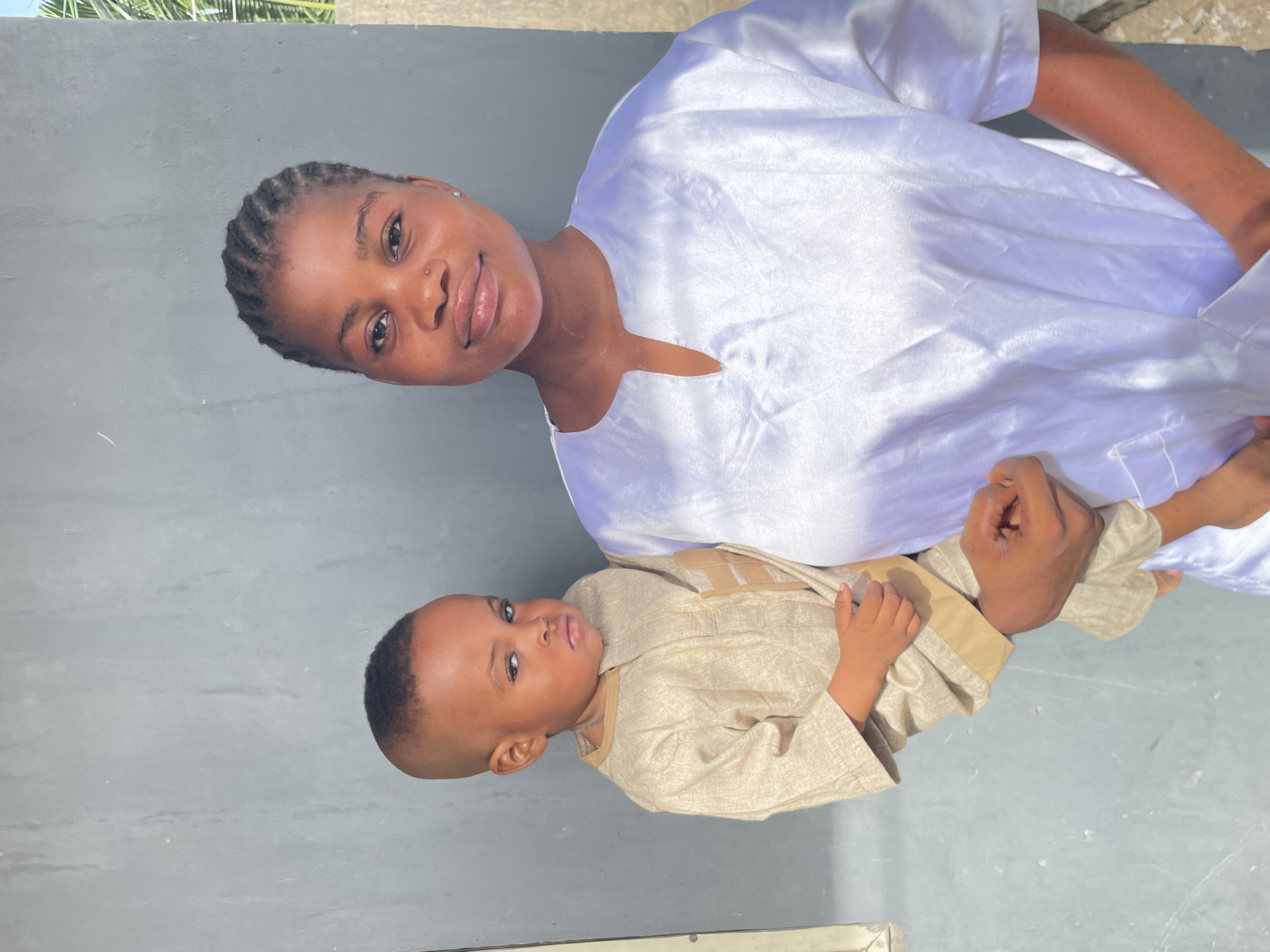 This portrait features a woman and a young child in an outdoor setting in Badagry, Nigeria. The woman is positioned on the left side of the frame, looking directly at the camera with a gentle, soft smile. She has dark skin, braided hair pulled back, a nose piercing on her left nostril, and a small earring in her left ear. She is wearing a luminous, white, long-sleeved garment with a distinct V-neck or split neckline, which appears to be made of a satin-like fabric.
She holds the hand of a young boy, who is situated slightly below and to her right. The boy has dark skin, a neat, short haircut, and gazes towards the camera with a neutral to slightly serious expression. He is dressed in a light beige or tan long-sleeved garment, possibly traditional attire, featuring a textured fabric and darker tan accents around the shoulders and chest.
The background consists of a plain light grey wall, which fills most of the upper and middle portions of the image. Along the very top edge of the frame, some green foliage, likely palm fronds, is visible, suggesting an outdoor environment. The lighting is bright and even, indicating daytime conditions. There is no visible text in the image. The overall mood is calm and personal.