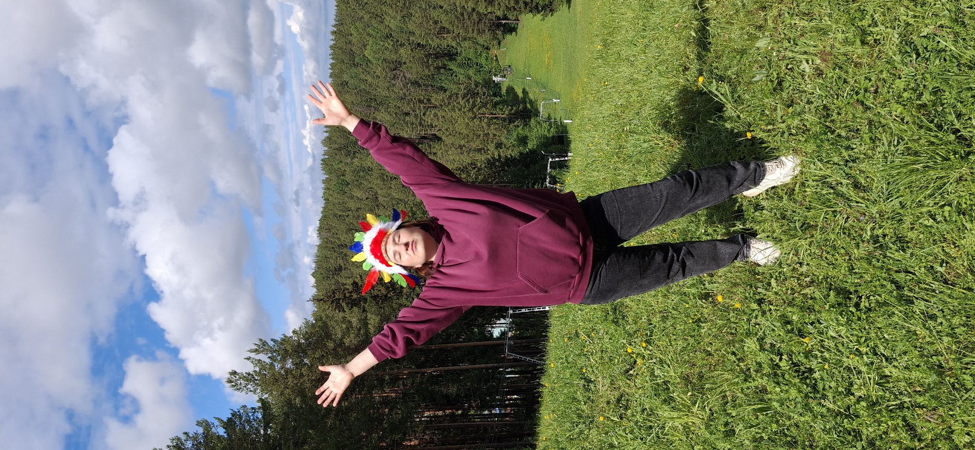 A young person, likely female, is lying on her back on a grassy field. She is wearing a burgundy hooded sweatshirt and black pants, along with white sneakers. On her head, she wears a feathered headdress, reminiscent of Native American attire. Her arms are outstretched on either side, as if making a snow angel.
The setting is outdoors in a grassy area with a forest backdrop. In the distance, there is a slight incline with more grass and possibly a ski lift structure. The sky above shows a mix of blue and white clouds. The image was taken in Прикамский, Russia.