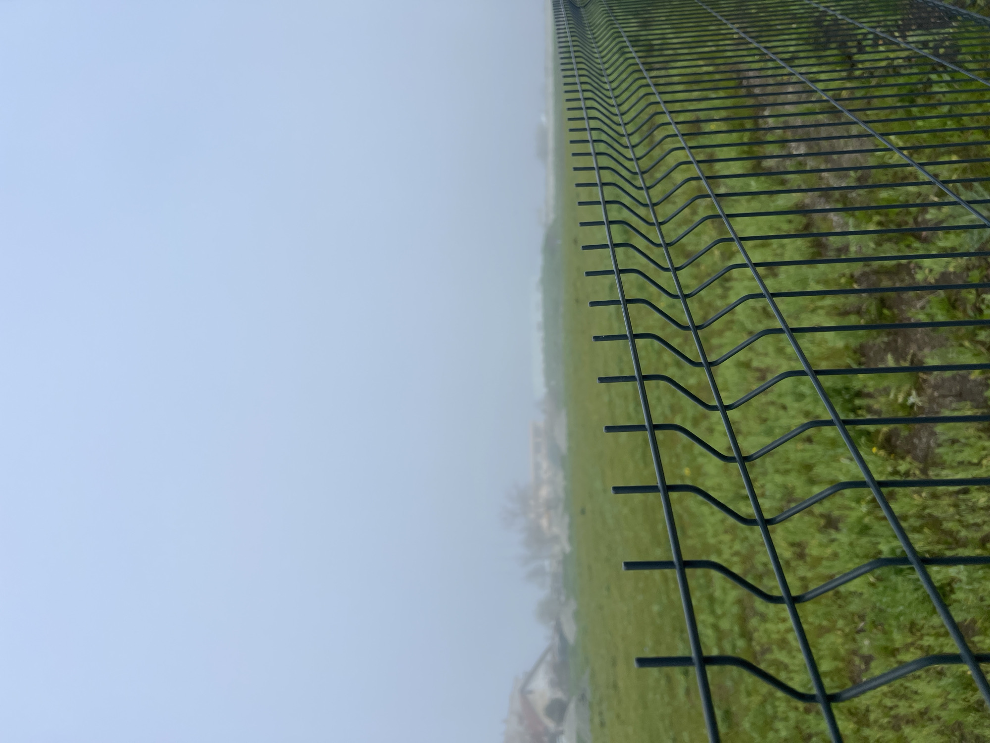 The photograph depicts a chain-link fence running vertically through the frame. To the right of the fence, there is green grass and some indistinct foliage or undergrowth. In the background, there appears to be some buildings or structures obscured by mist or fog. To the left of the fence, a dense fog or mist fills the scene, reducing visibility. The picture is taken at an angle, giving a perspective view along the length of the fence. The exact location within Burgas, Bulgaria, is not discernible from the visual information provided.