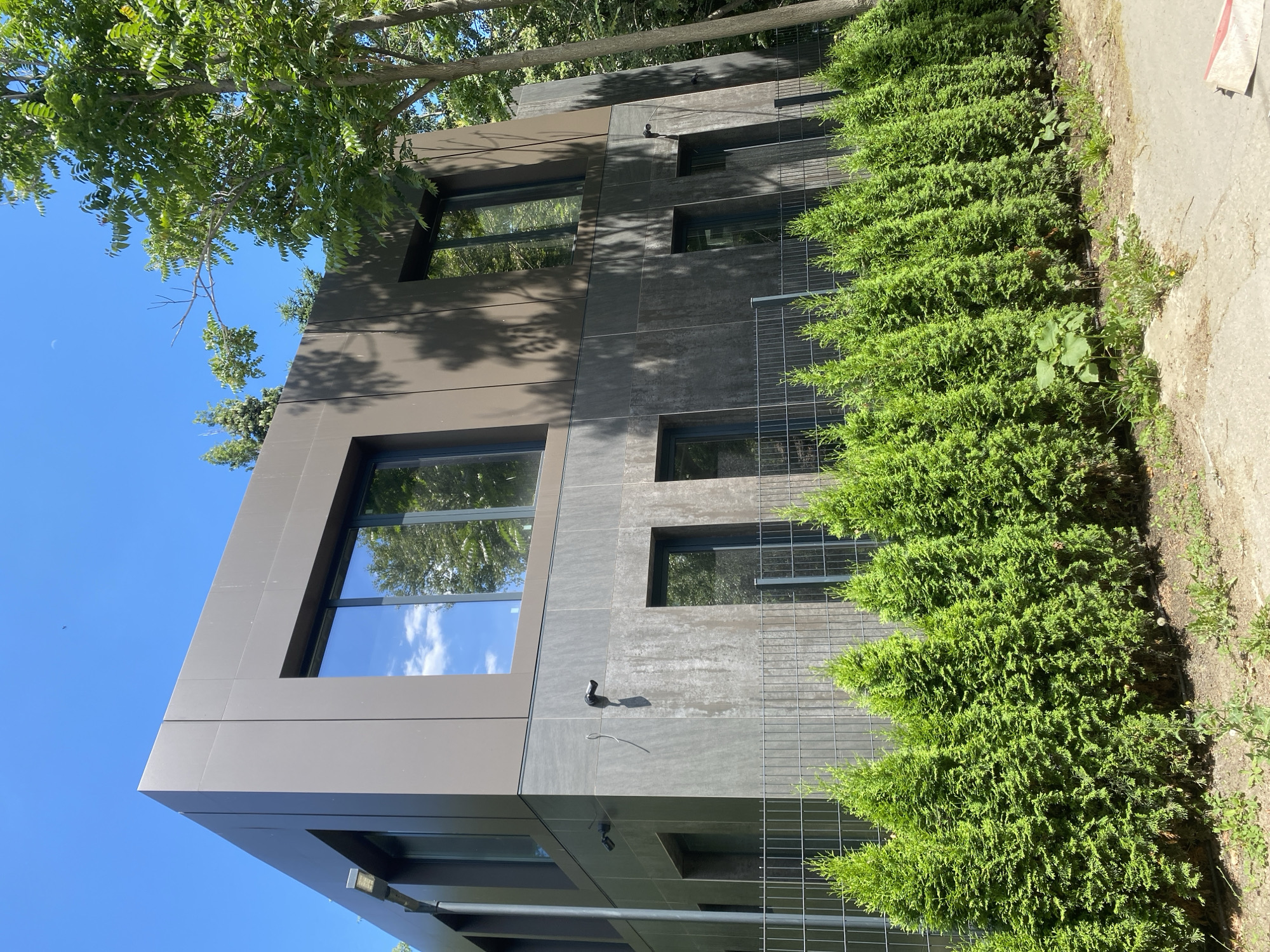 The image depicts a modern, two-story building with a dark brown facade on the upper level and a textured gray facade on the lower level. The building features large, dark-framed windows that reflect the blue sky and trees. A metal fence runs along the front of the property, behind a row of neatly trimmed, vibrant green evergreen bushes. Two visible security cameras are mounted on the lower facade. The surroundings are lush with green trees, casting shadows on the building. The sky is clear and bright blue. The ground in front of the bushes is dirt and some vegetation. The scene suggests a contemporary residential or commercial property. This image was taken in Varna, Bulgaria.