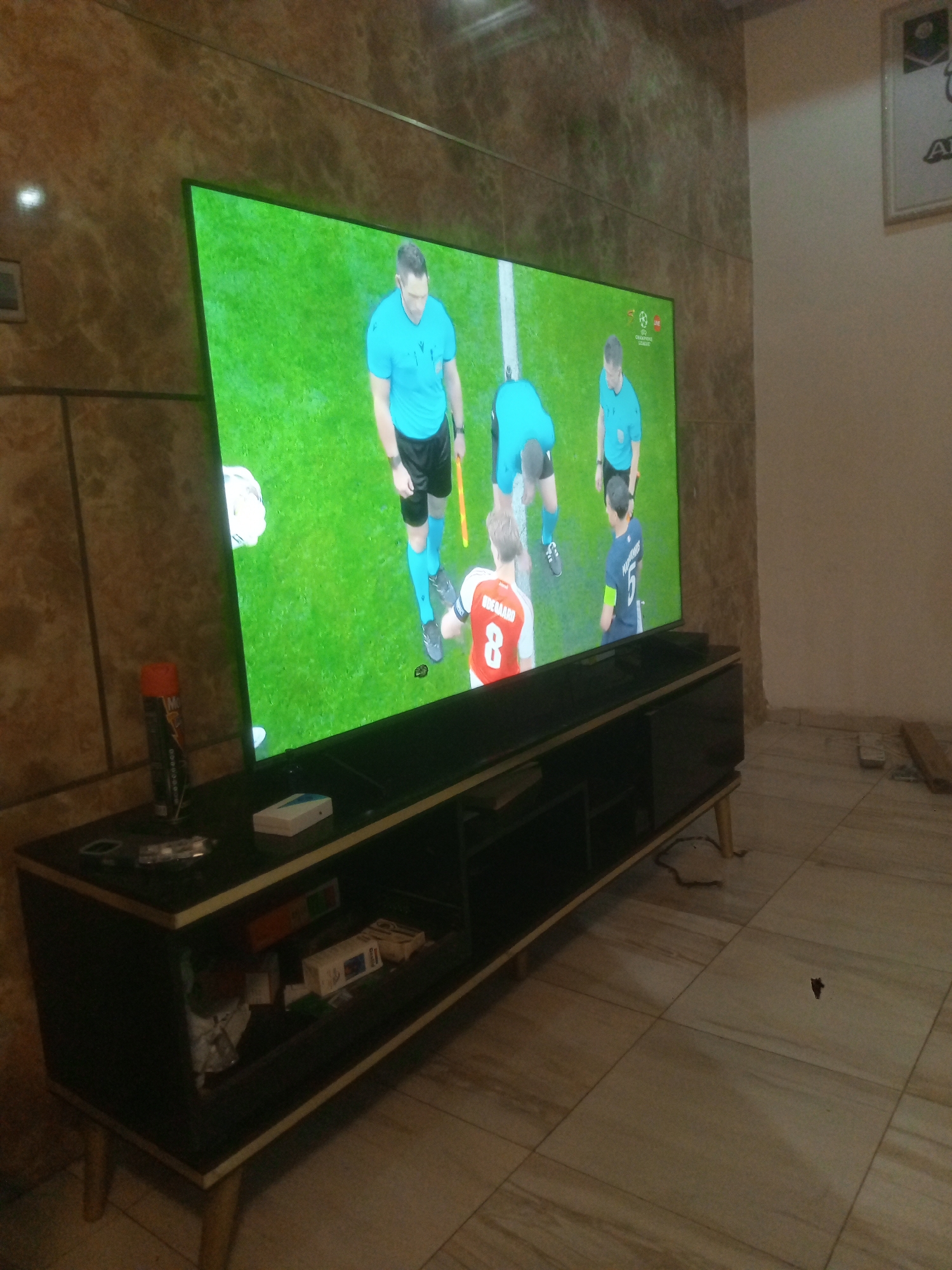 Here is a forensic description of the image:
The image shows a television displaying a soccer game, likely a high-profile match given the presence of referees and recognizable players. The background appears to be a living room or entertainment area. The TV is placed on a dark-colored media console with storage space.
**People:**
* There are multiple individuals on the television screen, including:
* Referees wearing bright blue uniforms with black shorts.
* Soccer players in team jerseys with identifiable numbers. One player is wearing a jersey with the number "8" and the name "ODEGAARD" on the back.
* Another player is wearing a jersey with the number "5" and the name "MARTINELLI" on the back.
**Subjects:**
* A soccer game is in progress on the television.
**Objects:**
* Television
* Media console: Dark color with light-colored legs
* Various items on the media console (e.g., a spray can, remote controls, white box)
* Items within the media console's storage compartments (boxes, cables, etc.)
* Tiled flooring
**Actions:**
* The main action is the display of a soccer game.
**Interactions:**
* The television is displaying the soccer game, which is the focal point.
**Environment:**
* Indoor setting, possibly a living room.
* Tiled flooring.
* The wall behind the TV has a textured pattern.
* Adjacent wall is plain and light colored.
* There is what looks like framed artwork on the wall.
**Location:** Unknown, Unknown. The image's metadata does not contain the location.