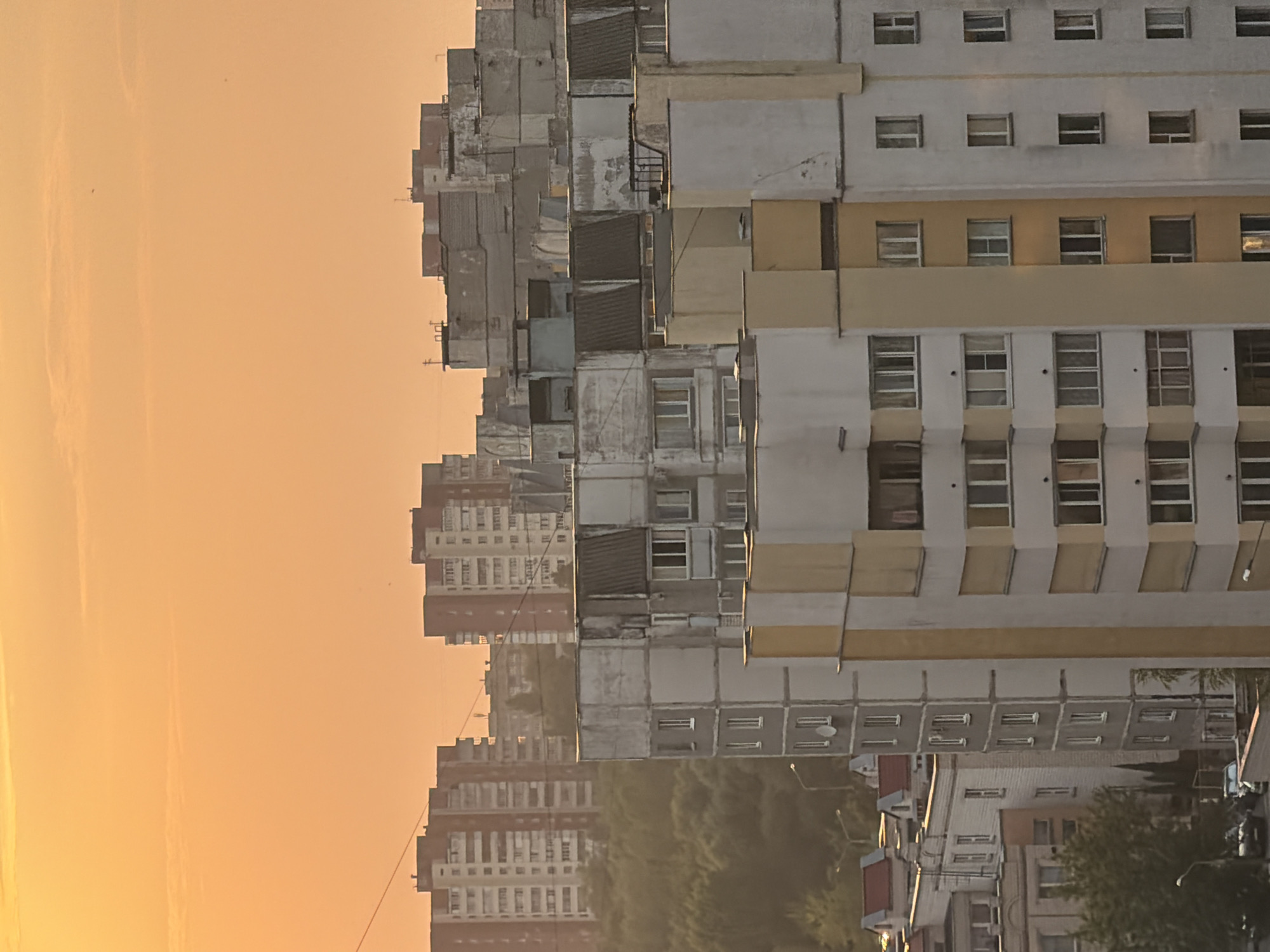 The image captures a cityscape of Homyel, Belarus, during what appears to be sunset or sunrise, given the warm orange hue of the sky. The foreground is dominated by the side view of a multi-story apartment building with a light grey exterior and vertical yellow accents. Numerous windows are visible, some with closed blinds, others appearing dark or with indistinct interior details.
Behind this building, a slightly older, more weathered apartment complex is visible, characterized by its grey concrete facade and varied window designs, some with visible balconies or extensions. Further in the background, more residential buildings of varying heights and architectural styles can be seen. The sky above is a uniform, soft orange, with faint horizontal cloud formations.
The scene depicts a densely populated urban environment with a focus on residential architecture. The presence of trees and some distant foliage suggests a degree of greenery within the city. No people are clearly visible in the image. The overall impression is one of a typical residential area in a Belarusian city during a period of low light.