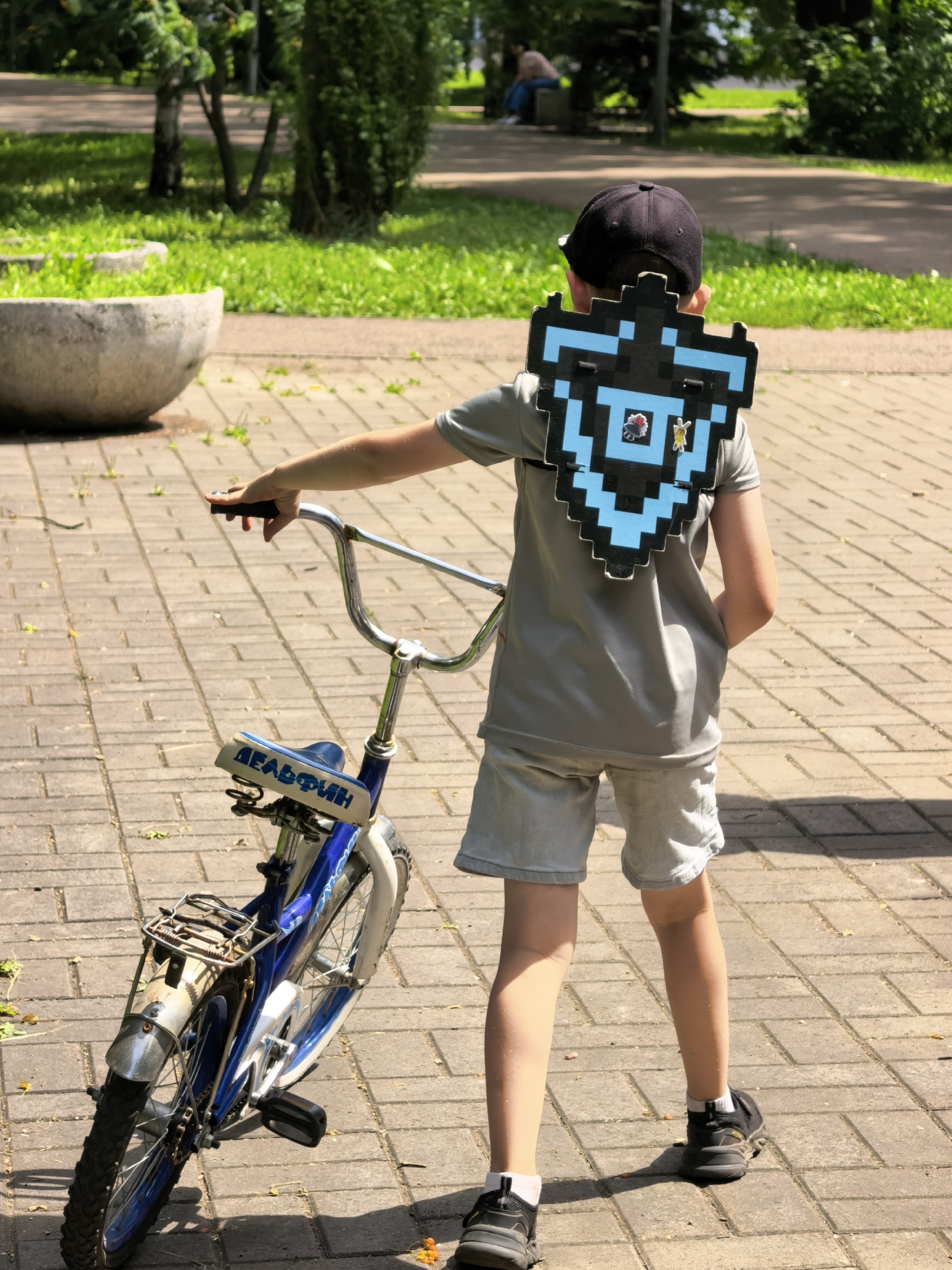 A boy, facing away from the camera, stands on a paved walkway in a park. He is wearing a grey t-shirt, light grey shorts, a black baseball cap, and dark sneakers. On his back, he has a pixelated, blue and black shield-like decoration. He is holding the handlebars of a small blue bicycle. The bicycle has a white saddle with blue Cyrillic lettering that reads "ДЕЛЬФИН" (DELFIN). The background shows green grass, trees, and in the distance, a person sitting on a bench. The lighting suggests it is daytime.