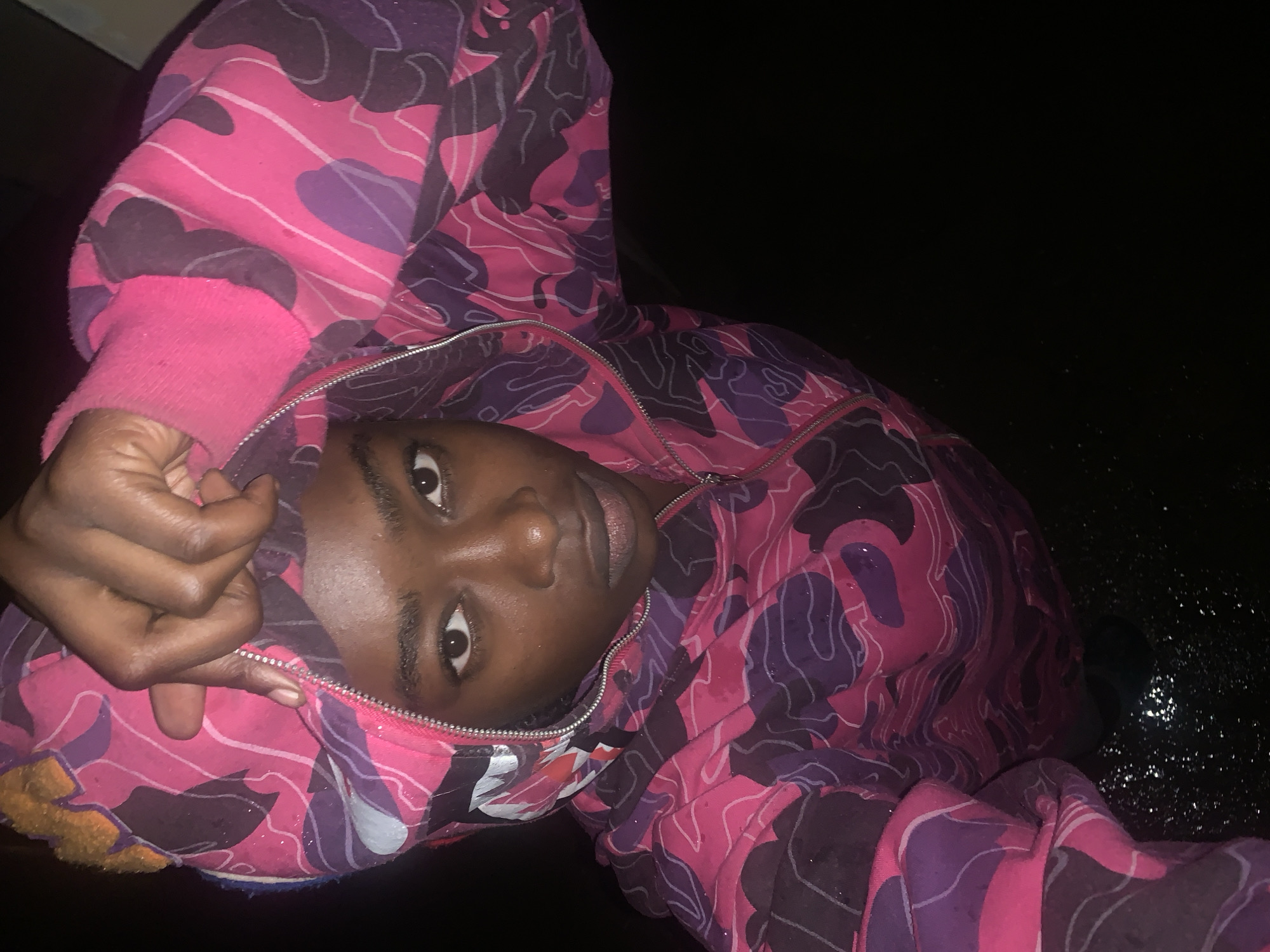 A portrait-style photograph, rotated counter-clockwise, depicts a young person, likely male, with dark skin, looking directly into the camera. The subject is wearing a full-zip hoodie with a bold camouflage pattern in bright pink, purple, and black. The design of the hoodie, particularly a visible shark face element near the zipper in one of the cropped images, suggests a style similar to "A Bathing Ape" (BAPE). The hoodie features solid pink cuffs.
The person's eyes are open, and their expression is neutral to slightly serious. One hand is visible, resting near the collar or hood of the jacket. The background is predominantly dark black, indicating a night-time setting or a very dimly lit environment. Small, reflective specks, possibly glitter, water droplets, or dust, are visible on the hoodie and in the dark background. The overall activity appears to be posing for a photograph. There is no visible text in the image. The location context for this media file is Rafin Par, Nigeria.