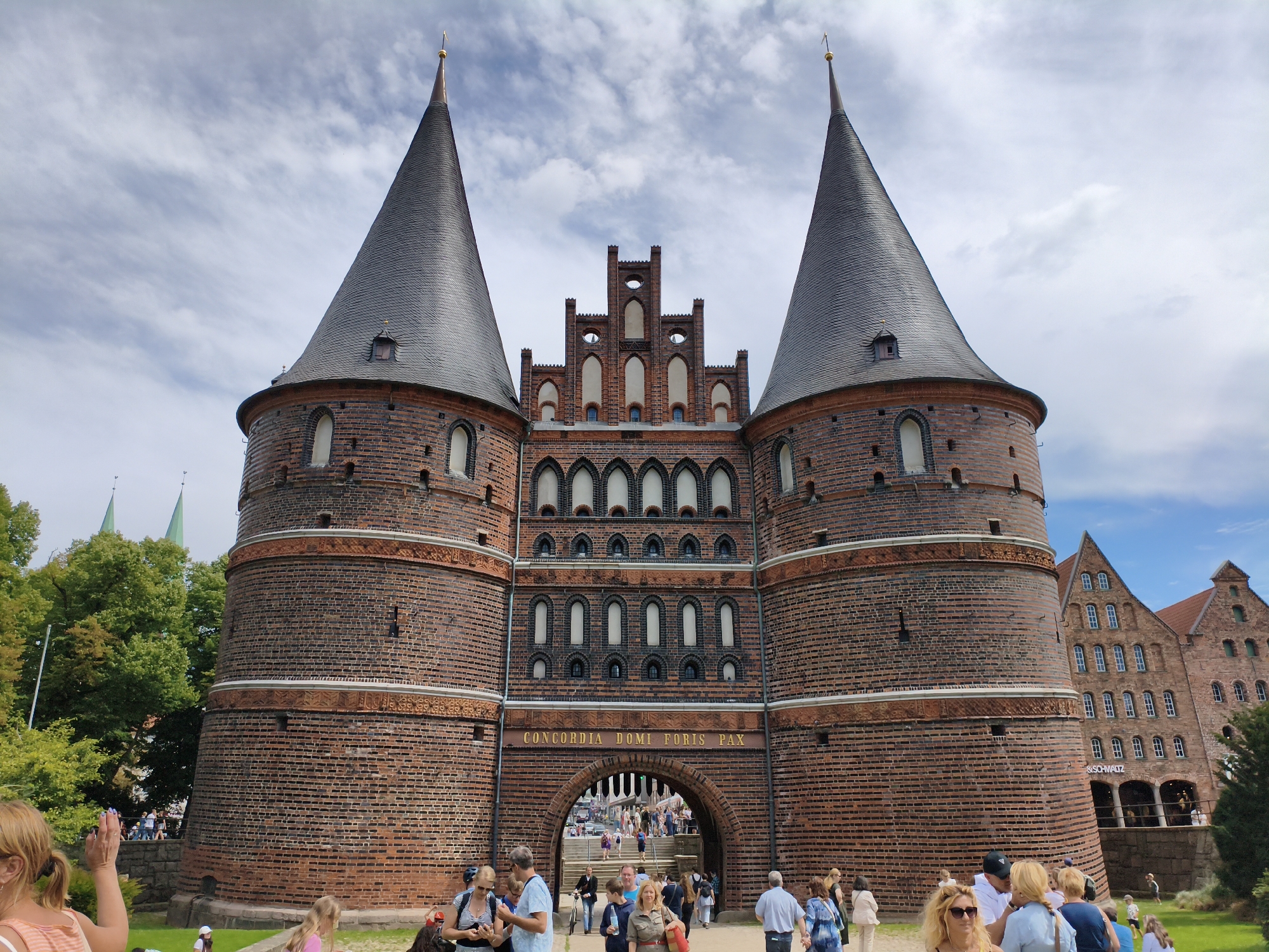 The image captures the Holsten Gate (Holstentor), a famous medieval city gate in Lübeck, Germany. The gate is a large, brick structure with two prominent conical towers and a central section featuring multiple levels of arched windows. Above the main archway, an inscription reads "CONCORDIA DOMI FORIS PAX." The sky is partly cloudy. In the foreground, a diverse group of people are gathered, some walking and others standing. A woman with blonde hair and a striped top is in the left foreground, raising her hand. To her right, other individuals are visible, including a man in a blue shirt and a woman in a green jacket. People are seen walking through the archway of the gate, indicating it is an active thoroughfare. To the left of the gate, lush green trees and the spires of other buildings are visible. To the right, a traditional brick building with multiple windows stands adjacent to the gate. The scene appears to be a public space on a pleasant day, with tourists and locals interacting and exploring the historic landmark.