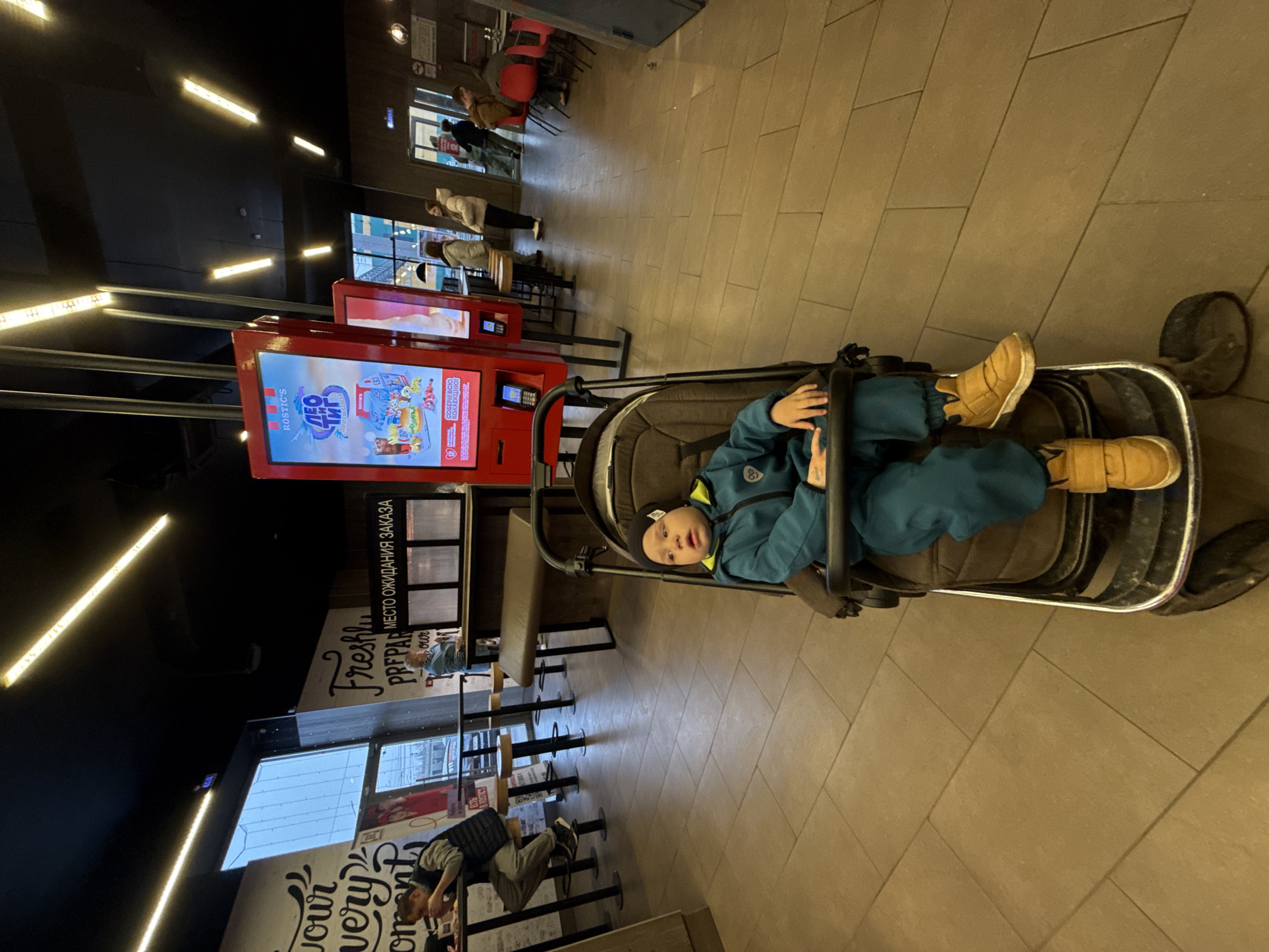 The image shows a child lying in a stroller inside a Rostik's restaurant, a fast-food chain formerly known as KFC, located in Rusanovo, Russia. The child is wearing a blue jacket and tan boots. The stroller is parked near a self-service kiosk advertising the "Leo" collection and other menu items. Other patrons can be seen standing in line and sitting at tables in the background. The restaurant has a modern interior with dark ceilings, bright lighting, and tile flooring.