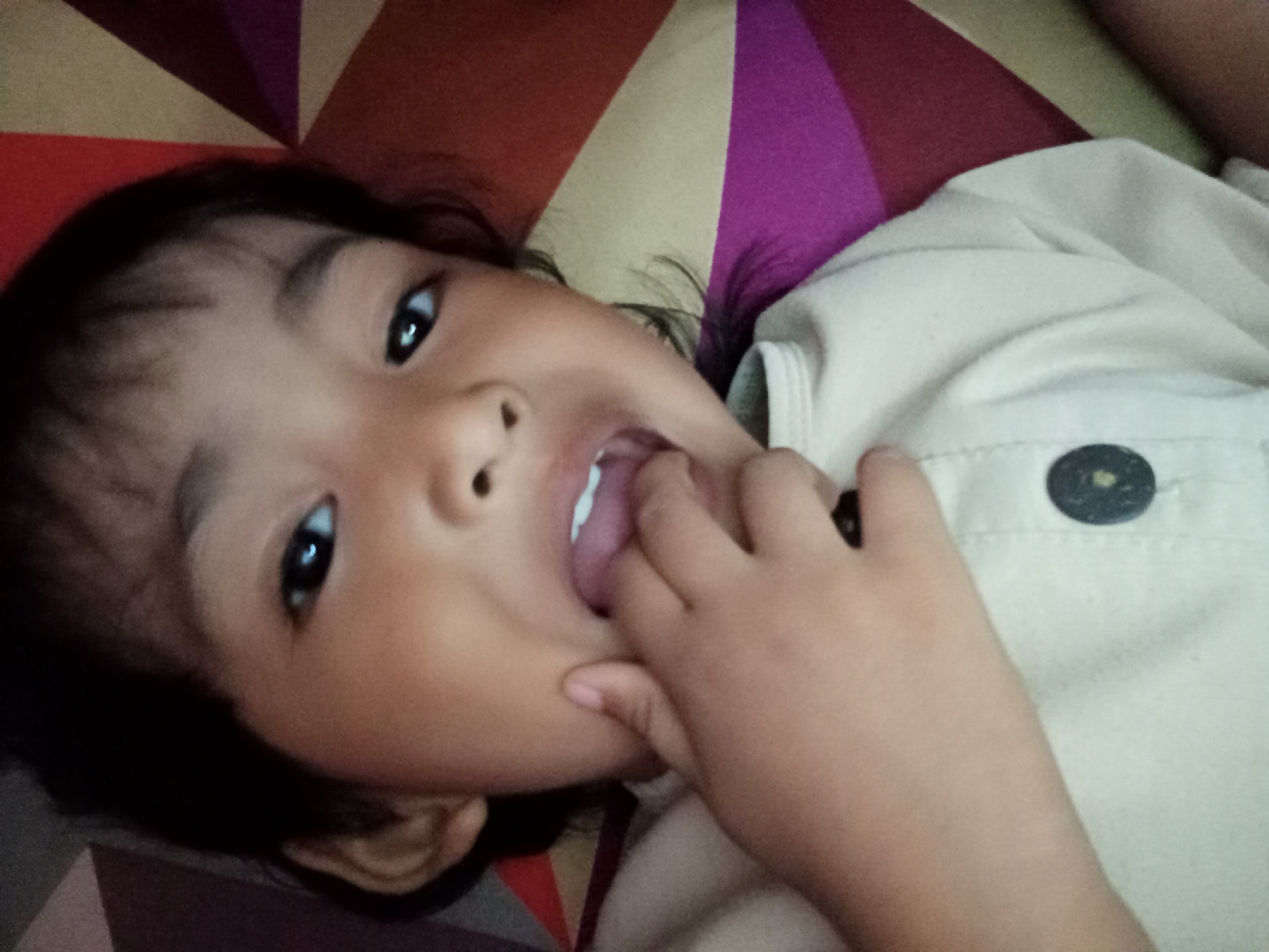 The image shows a close-up of a young child lying on their back on a patterned surface. The child is looking towards the camera with wide, dark eyes, and has their finger in their mouth, with their tongue visible. Their mouth is slightly open. The child is wearing a light-colored shirt with a dark button. The patterned surface appears to be a blanket or bedspread with geometric shapes in shades of red, yellow, purple, and possibly brown. The lighting suggests an indoor setting, potentially during the daytime given the brightness. The overall mood appears playful or curious due to the child's expression and gesture. There is no visible text in the image. The location context is Bloro, Indonesia.