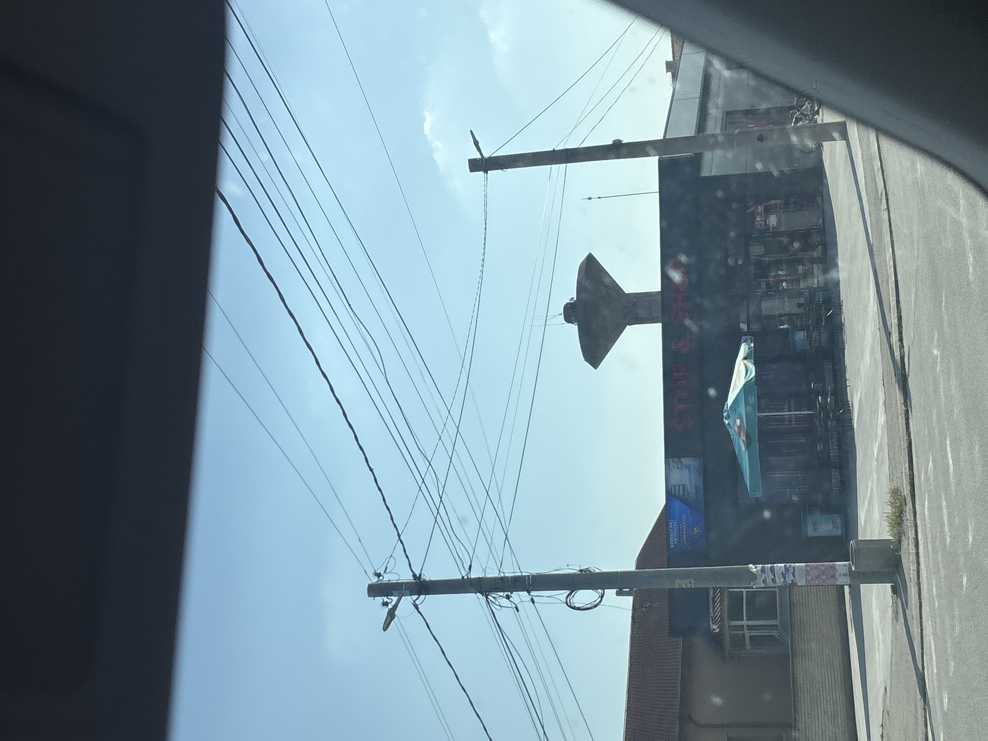 The image depicts a daytime street scene in Rakovski, Bulgaria, captured from inside a vehicle. The sky is clear blue with sparse white clouds. Two concrete utility poles dominate the midground, heavily laden with numerous overhead electrical and communication wires that crisscross the frame. The left pole features a street light arm and has remnants of paper posters and a coiled cable bundle near its base, where a concrete waste receptacle or planter is situated on a patch of dry grass.
To the right of the left pole stands a dark-fronted, single-story commercial building, with a prominent sign reading "STOP & SHOP" (partially visible as "STOP & SH"). In front of this building, an outdoor seating area is visible, complete with a blue and white striped umbrella, tables, and chairs. A blue advertising banner, possibly with Cyrillic text, is mounted above the building's entrance. A motorcycle or scooter is parked near the right side of this building.
Behind the left utility pole and adjacent to the commercial building, a residential-style building with a reddish-brown tiled roof and a distinctive bay window section is visible. A low brick wall runs along the front of this property. In the background, rising above the commercial building, a concrete water tower with a unique inverted conical top section on a cylindrical base stands prominently against the sky. The street in the foreground is asphalt, showing faint road markings. No individuals are visible in the scene.