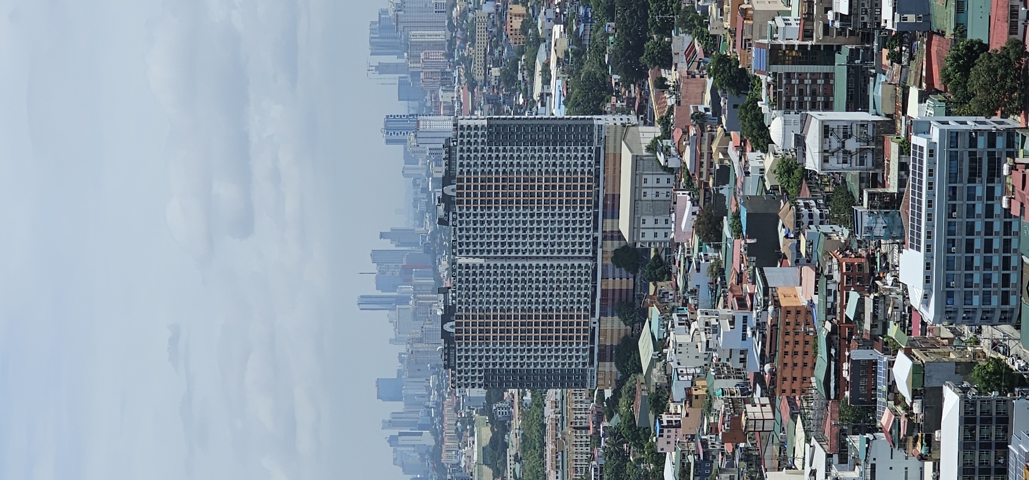 This is a medium-altitude, vertical, eye-level, full shot of a dense urban landscape in Makati, Philippines. The image is dominated by buildings of varying heights and architectural styles. In the foreground and mid-ground, there are numerous smaller residential and commercial buildings with colorful roofs and balconies. Many of these structures appear to be low-rise or mid-rise, creating a complex tapestry of urban sprawl.
A prominent feature in the mid-ground is a very large, modern, dual-tower residential building with a repetitive grid pattern of balconies and windows. The building has a light gray facade with alternating bands of orange and brown on its upper sections. To the left of this large building, there is a sprawling, low-rise structure with a light-colored exterior and numerous windows.
In the far background, a distant skyline of taller, modern skyscrapers is visible, suggesting the presence of a central business district. These buildings are hazy due to atmospheric conditions. The sky above is overcast with light gray clouds, obscuring any direct sunlight. The overall impression is of a vibrant, densely populated city with a mix of old and new construction. There are no people or vehicles clearly identifiable in the image. The environment appears to be built-up and developed, with limited visible green spaces apart from scattered trees.