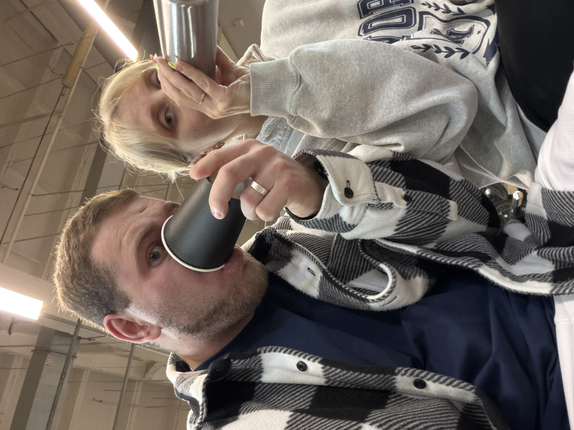 Two individuals are positioned closely together, appearing in a low-angle, candid photograph. The foreground features a man drinking from a black paper cup; he is wearing a navy blue shirt underneath a black and white plaid flannel jacket, and a wedding band is visible on his left ring finger. Behind him, a woman is holding a silver thermos, visible near her face. She wears a light gray sweatshirt with dark blue designs. The setting includes an industrial ceiling with visible fluorescent lighting, potentially suggesting a retail or commercial space. This photograph does not give any information of it being taken in the city of Chaikovsky, Russia.