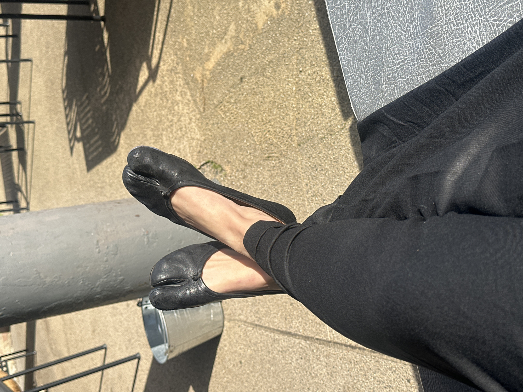 A first-person, downward-angle photograph captures a person's lower legs and feet, crossed at the ankles, in an outdoor setting. The subject wears black trousers or leggings and black leather split-toe (Tabi-style) flat shoes. Bright, direct sunlight illuminates the scene, casting distinct, elongated shadows, primarily from thin-framed objects consistent with outdoor furniture, across the light-colored, granular paved ground. In the mid-ground, to the left, a grey cylindrical pillar stands beside a silver metallic bucket. The person appears to be seated, with a portion of a grey, textured surface, possibly a bench or table, visible in the lower right foreground. The scene depicts an outdoor urban environment, consistent with a public area or cafe in the City of London, United Kingdom.