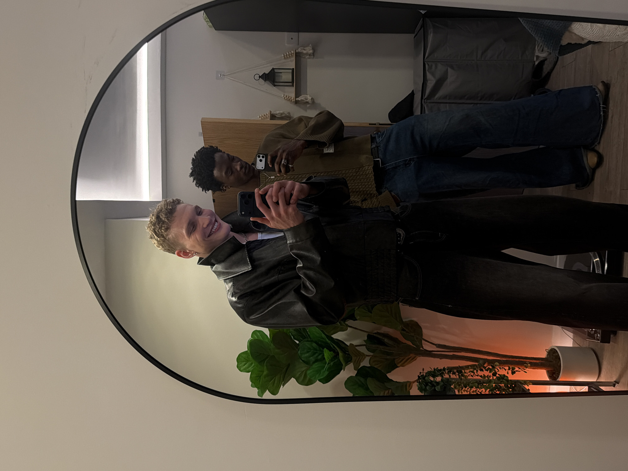 The image, taken in London, United Kingdom, shows a mirror selfie of two young men captured in a large, arched mirror with a black frame. The indoor setting features white walls and a light wooden floor.
In the foreground of the reflection, a man with light, curly hair smiles widely at the camera, holding a smartphone with both hands to take the picture. He wears a black leather jacket over a white top. Behind him, further in the reflection, stands another man with dark, curly hair, sporting a dark olive-green knitted sweater, blue jeans, and a ring on his finger. He is also holding a smartphone, seemingly engaged with its screen.
The room's decor includes a light wooden furniture piece or headboard behind the second man. Above it, a decorative wall hanging features a small black lantern and white tassels. To the right side of the reflection, a large potted houseplant with broad green leaves, resembling a fiddle-leaf fig, stands alongside a smaller plant. A warm, orange light illuminates the wall behind these plants, casting a gentle glow. A dark grey storage bag or box is visible on the floor behind the man in jeans. A narrow vertical light strip illuminates a white wall to the far left of the reflection.
The scene suggests an evening or nighttime setting, relying on artificial lighting for illumination. The prominent smile of the man in the foreground conveys a positive and relaxed mood during this casual mirror photo session. No discernible text is visible within the image.