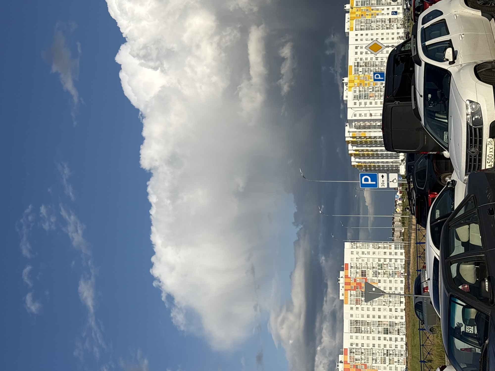 The image shows a view of apartment buildings in the city of Alexandrovka, Russia, under a dramatic sky filled with large clouds. In the foreground, several cars are parked, including a white SUV and a dark sedan. In the midground, there is a row of parked cars and a black van, with streetlights and parking signs visible. The apartment buildings, which are white with colorful accents in red, orange, and yellow, dominate the background. The sky features a mix of bright blue and dark, heavy clouds, suggesting a change in weather. The overall scene depicts an urban environment with residential buildings and a parking lot, set against a striking natural backdrop of clouds.