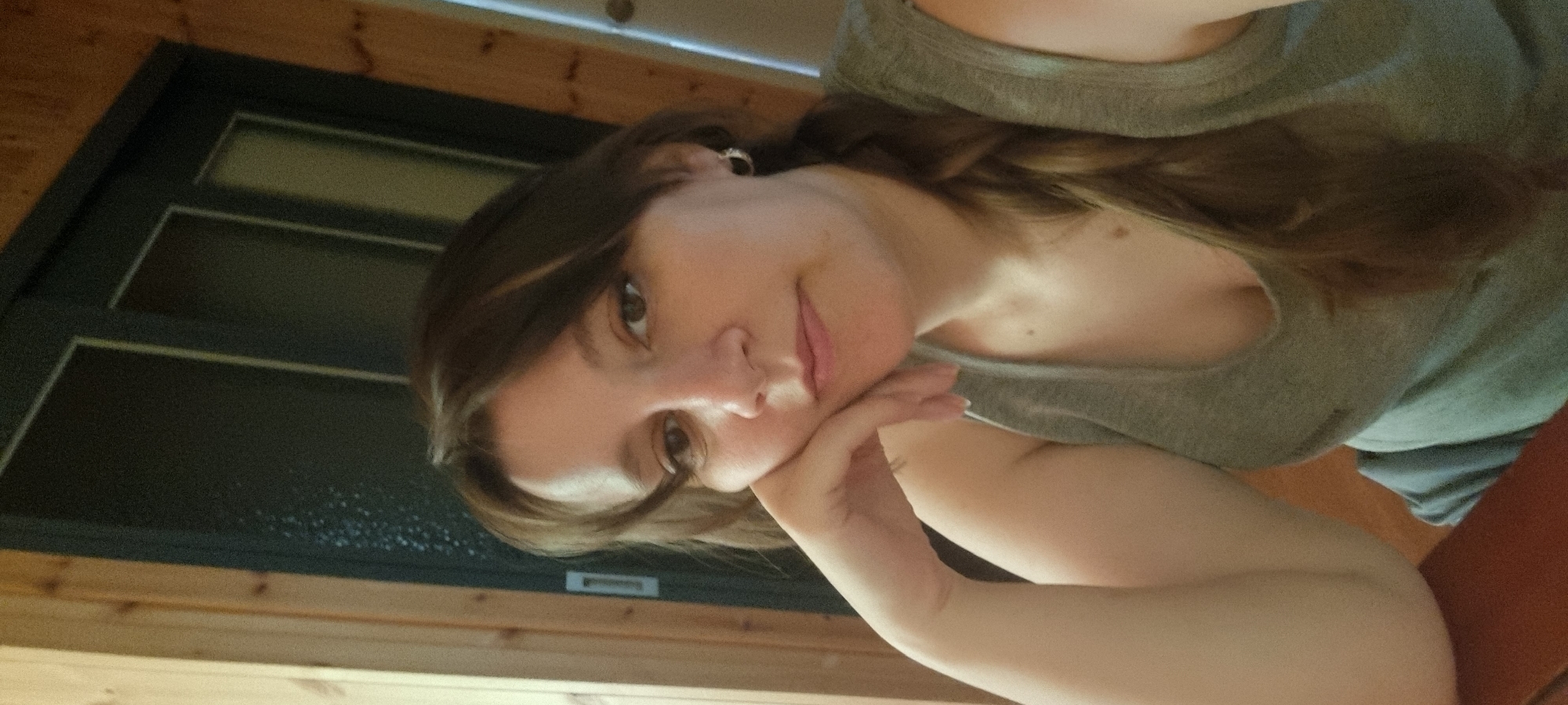 A close-up portrait captures a young woman in an indoor setting. The woman, likely in her 20s or 30s, has long brown hair styled with a visible braid over her left shoulder. She wears a plain grey tank top and a single earring in her visible left ear. Looking directly at the camera, she displays a subtle, gentle smile, with her left hand resting casually against her chin or cheek.
The background features warm-toned wooden panels, suggesting an interior wall or ceiling. To the left, a dark-framed structure, resembling a window or door with multiple glass panes, is prominent. The lower section of this glass appears to have small droplets or condensation on its surface. The lighting is soft and even, indicating a daytime indoor scene. A small portion of a brown or orange object, possibly a piece of furniture, is visible in the bottom right corner.
The image conveys a calm and relaxed atmosphere. No visible text is present. The scene is situated in Dunnae-myeon, South Korea.