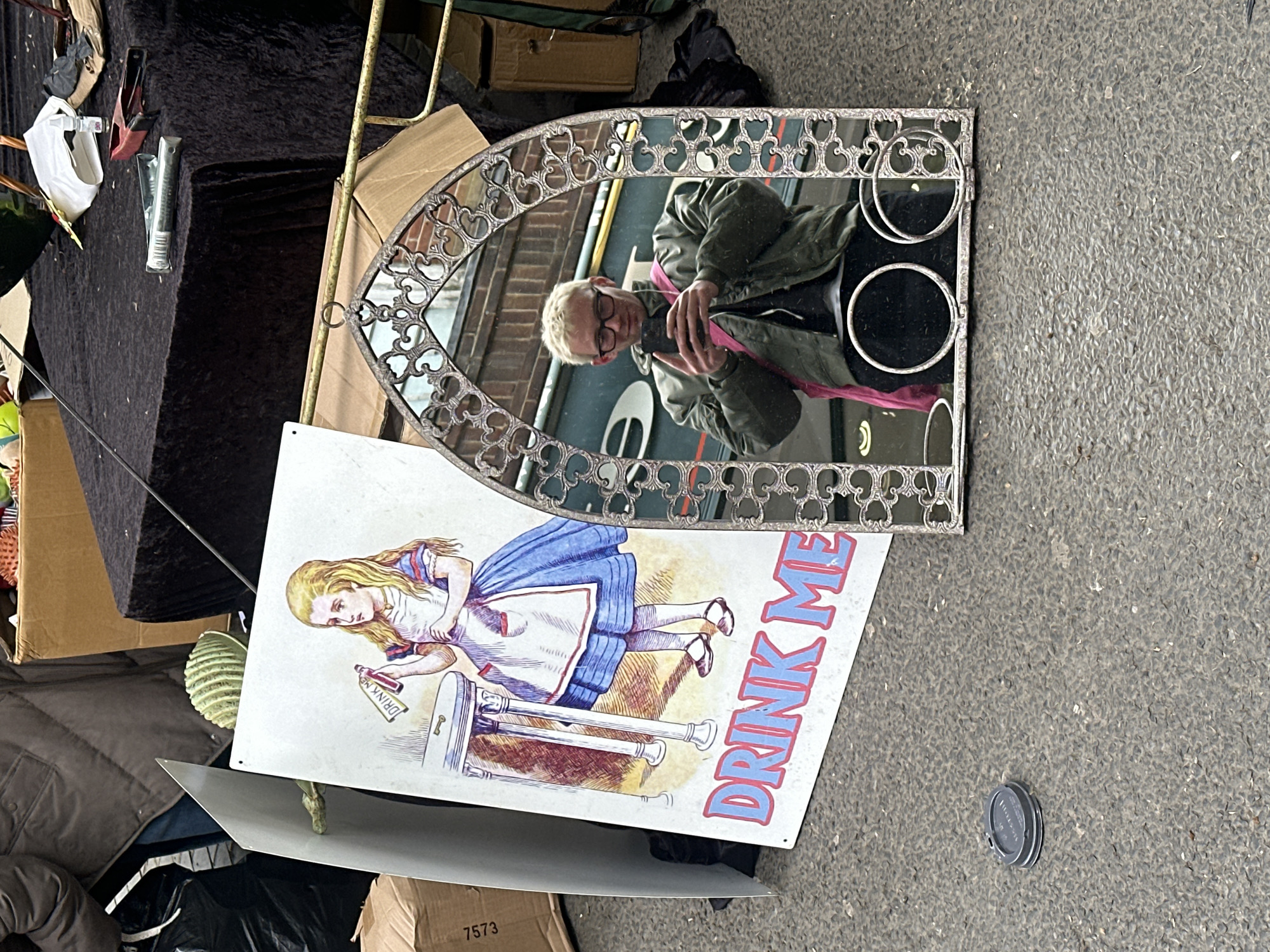 The image depicts a collection of objects strewn on a pavement or street in St Albans, United Kingdom. A mirror, reflecting a person taking a selfie, lies at an angle. The mirror has a silver, ornate frame. A white sign with an illustration of Alice from Alice in Wonderland and the text "DRINK ME" is also present. The scene appears to be part of an outdoor market or sale, with other items like boxes, a black draped table, and other unidentified objects scattered around. The ground is asphalt. A discarded can lid is visible on the asphalt.