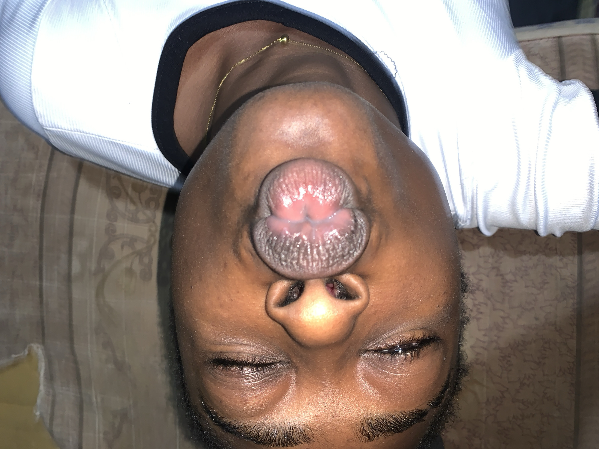A close-up, vertical, low-angle shot shows the face of a young person lying down, their head tilted back. Their eyes are closed, and their mouth is pursed, with their tongue sticking out and slightly curled. The person is wearing a white, ribbed t-shirt with a black trim around the neckline, and a thin gold chain with a small ball pendant is visible around their neck. The background consists of a patterned surface, possibly a bed or sofa. The image appears to have been taken indoors, given the lighting and the soft, textured background. There is no discernible information within the image to suggest a specific location within Rafin Par, Nigeria.