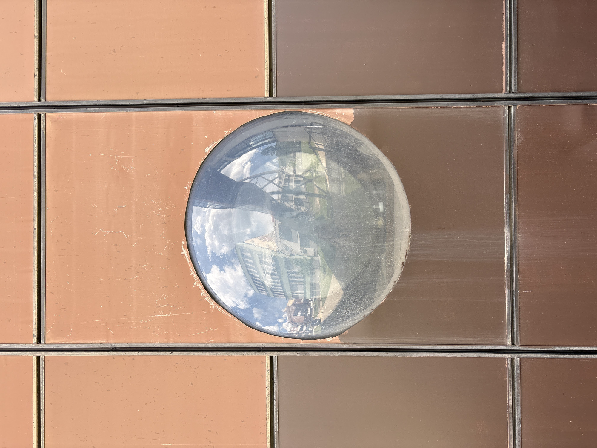 The image shows a close-up of an architectural detail. There are several rectangular brown panels arranged in a grid-like pattern, separated by dark-colored dividers. In the center of the grid is an oval window, reflecting the exterior environment. The reflection shows a building with a ladder and the sky with clouds. This window is on the facade of the Old Foundry Museum (Stara livnica), a historical museum in Kragujevac, Serbia.