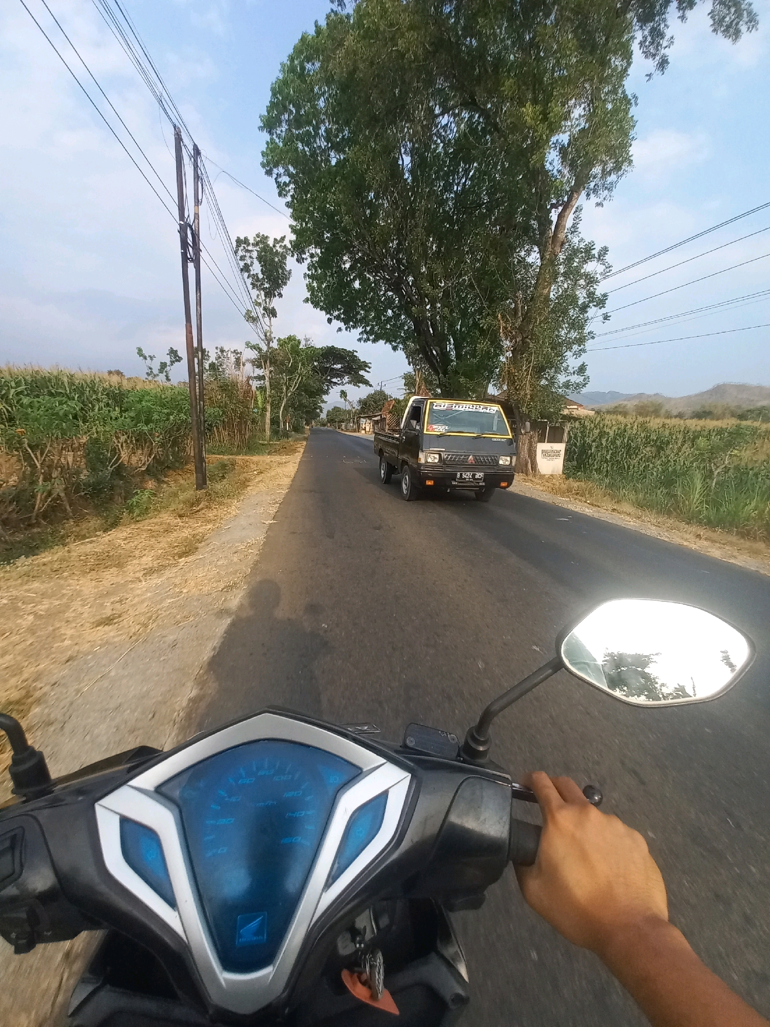 A first-person perspective shot shows a person riding a motorcycle on a paved road in a rural area. The motorcycle's blue-lit instrument cluster is visible in the foreground, with a hand gripping the right handlebar. The road extends into the distance, flanked by fields of green crops and trees. A black pickup truck with yellow accents is driving on the road ahead. Utility poles and power lines are present along the side of the road. The sky is a clear blue with scattered clouds. The scene appears to be in Ngarjosari, Indonesia, based on the context provided.