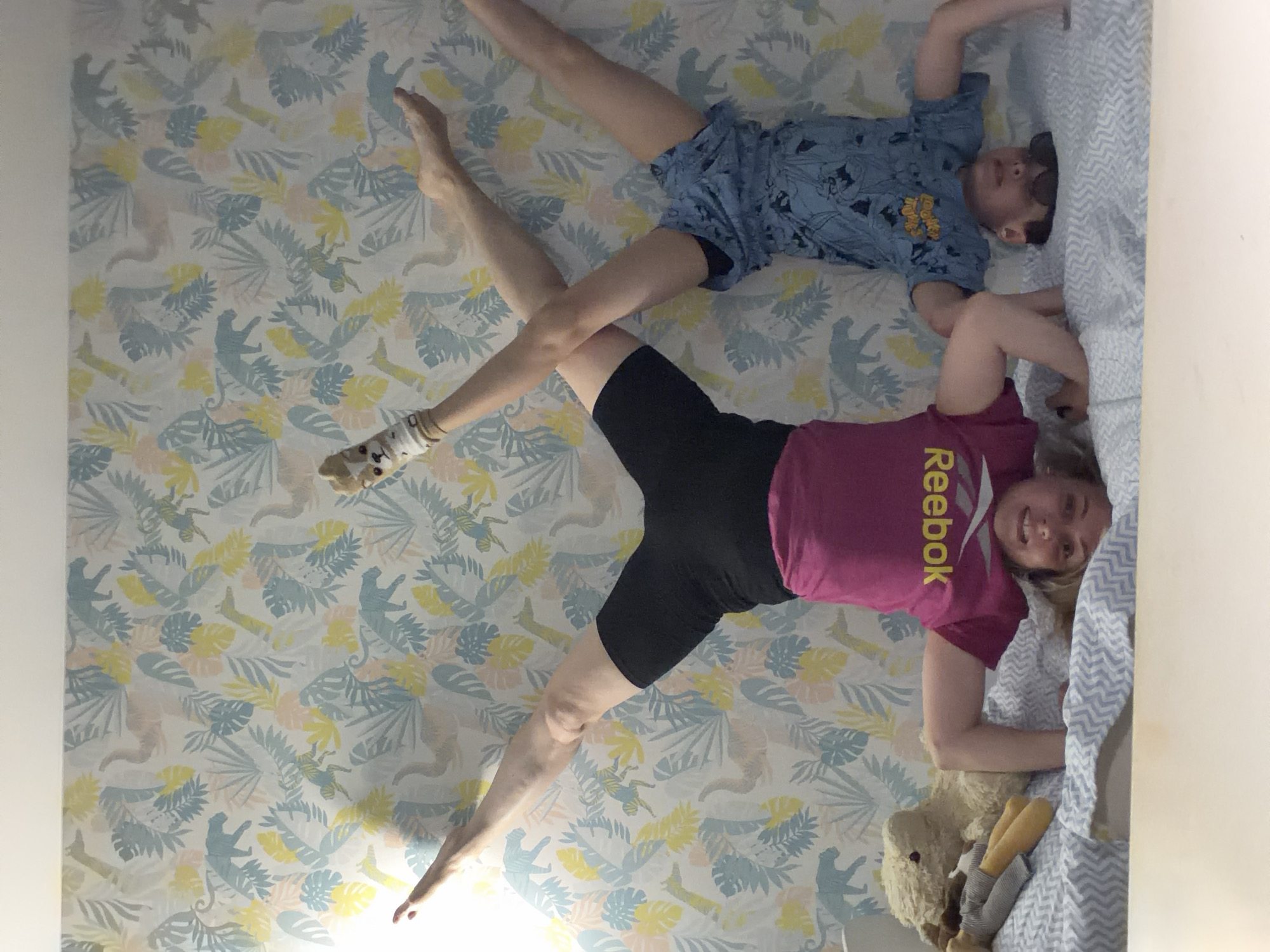 The image captures two individuals, likely a mother and child, playfully posing in what appears to be a bedroom in Votkinsk, Russia. The photo is rotated 90 degrees, creating an illusion that they are lying on a vertical surface or wall, though they are actually on a bed.
The adult female, with blonde hair, is wearing a dark pink t-shirt with the word "Reebok" printed in yellow, and black shorts. She is smiling broadly and looking towards the camera, her body positioned on the bed with her legs extended upwards. One foot wears a light-colored sock with a cartoon animal face.
Next to her, a young boy with short dark hair is visible, wearing a blue patterned short-sleeved top and blue shorts. He is also lying on his back, looking towards the camera with a smile, and his legs are extended and crossed with the adult's. He appears to have dark sunglasses pushed up onto his head or forehead.
The bed is covered with a patterned duvet or sheet featuring light blue, yellow, and pale pink leaves and stylized animal shapes, consistent with a whimsical or child-friendly theme. This pattern is also visible on the adjacent wall, suggesting matching decor. A blue and white zig-zag patterned pillow or bedding is visible under the adult female's head. A light brown teddy bear with yellow legs rests on the bed near the adult's lower body.
The scene conveys a joyful and playful moment between the two individuals. The lighting suggests it is daytime, with soft illumination.