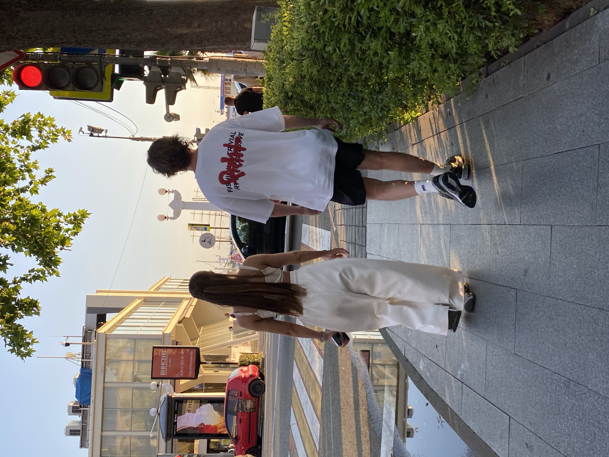 Two individuals, a man and a woman, walk away from the camera on a paved sidewalk. The woman is on the left, wearing a white crop top and loose white pants. She has long, dark hair pulled back. The man is on the right, wearing a white t-shirt with red lettering on the back and black shorts. He has medium-length, curly brown hair. They are walking on a sidewalk bordering a street with a pedestrian crossing. A red car is visible on the left side of the street. In the background, there are buildings, trees, and a traffic light with a red signal illuminated. The scene appears to be in a city, likely Sochi, Russia, given the context provided. The lighting suggests it is late afternoon or early evening, with sunlight casting long shadows.