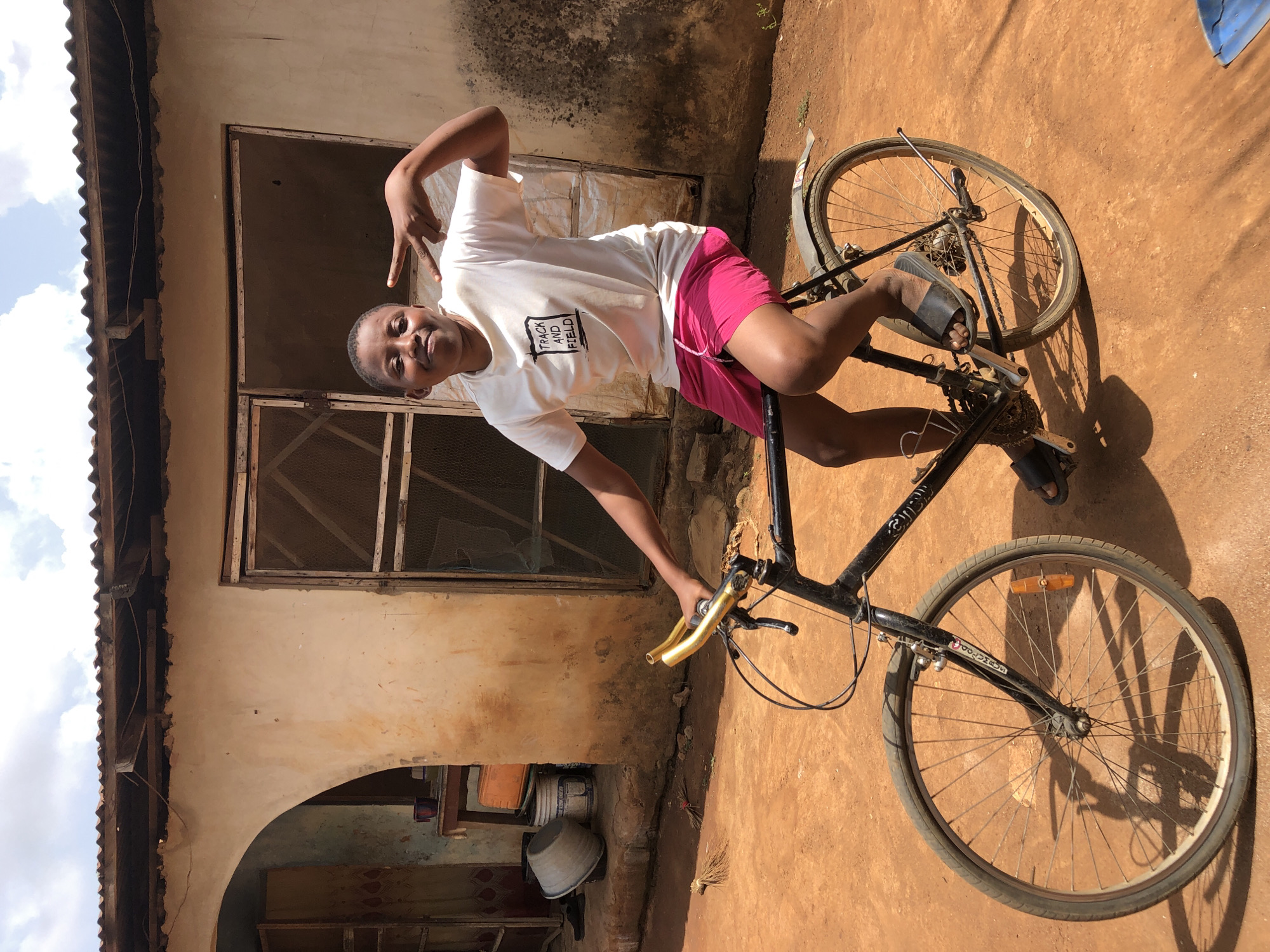 A person is straddling a black bicycle in front of a building with a rusted corrugated metal roof. The individual is wearing a white t-shirt with the words "TRACK AND FIELD" printed on it and pink shorts. They are also wearing black sandals. The bicycle has gold-colored handlebars. The building behind them has an arched opening with what appears to be a window with a metal grill. The ground is dirt, and there is a bucket visible in the background. The location is Unknown, Unknown. The person is posing and making a hand gesture.