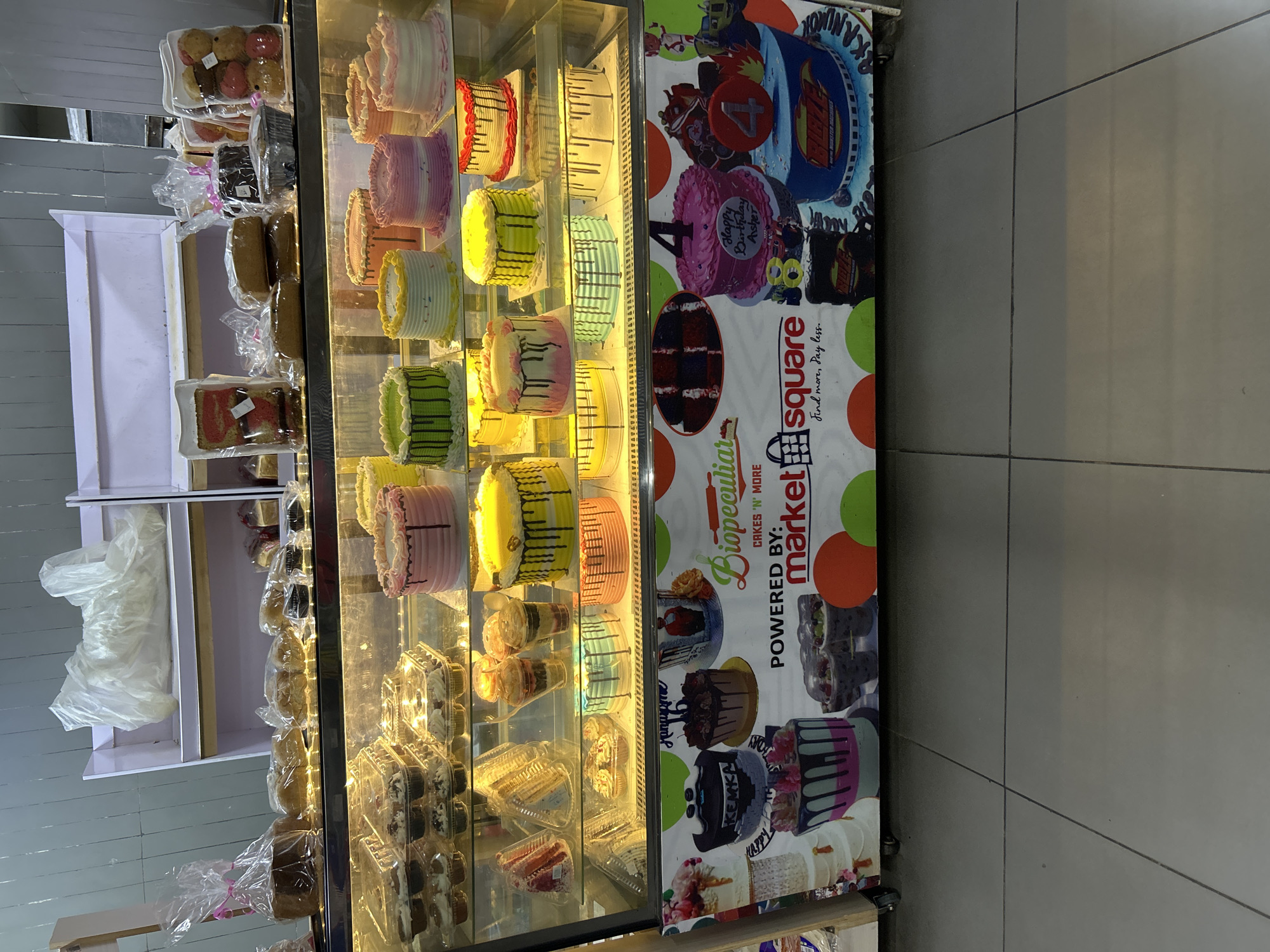 This is a photograph of a refrigerated display case filled with various cakes and baked goods. The display case is tall and appears to be made of glass, allowing a clear view of the items inside. On the left side of the display, there are shelves containing what appear to be individually packaged pastries or small cakes, some wrapped in plastic.
The main focus of the display is on the right side, where a variety of colorful cakes are arranged. These cakes are round and layered, decorated with frosting in different colors like pink, yellow, green, and blue. Some cakes have intricate patterns or drizzles of frosting. There are also smaller cupcakes or mini-cakes in clear plastic containers on the lower shelves.
A prominent graphic banner is attached to the side of the display case. This banner features bold text and colorful illustrations of cakes. Visible text on the banner includes "Biopeculiar," "CAKES 'N' MORE," "POWERED BY: market square," and "Find more, Pay less." There are also images of cartoon characters and birthday-themed graphics, including one that says "Happy Birthday Asher."
The background of the image shows a tiled floor and a portion of a wall. The lighting within the display case is bright, illuminating the baked goods. The overall impression is that of a bakery or a section of a store dedicated to selling cakes and desserts. There are no people visible in the image. The activity depicted is the display of food items for sale. The time of day and weather are not discernible from the image. The setting appears to be indoors.