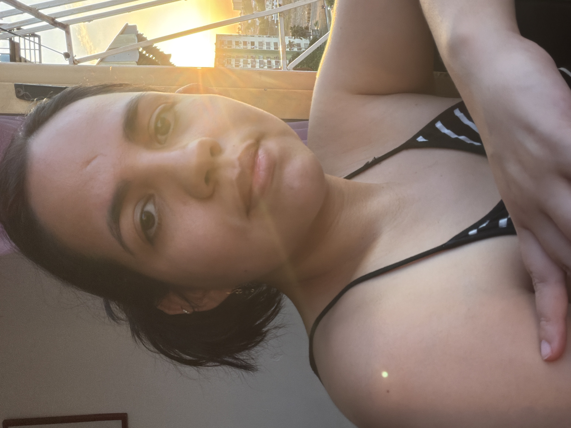 A woman with dark hair, wearing a black and white striped bikini top, poses for a close-up selfie. The background shows a cityscape under a bright sunset. The location is identified as Ciudad Guayana, Venezuela based on visible architectural features.