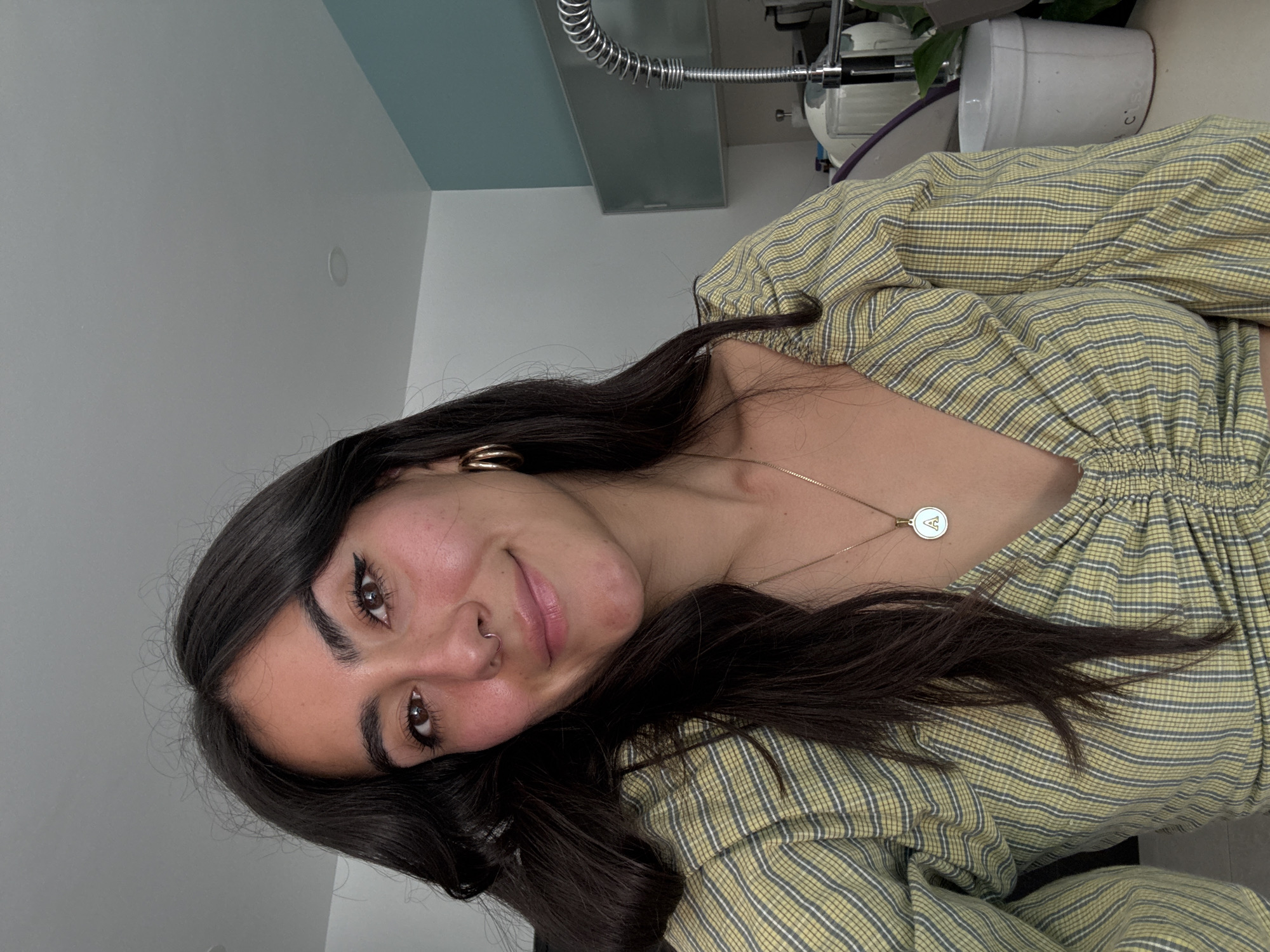 A young woman with dark hair and brown eyes poses indoors. She has a nose ring and is wearing gold hoop earrings and a gold necklace with a circular pendant. Her top is light green and yellow plaid. The background includes a white wall, a blue ceiling accent, a metal spring coil, and a white container. The location is not specified.