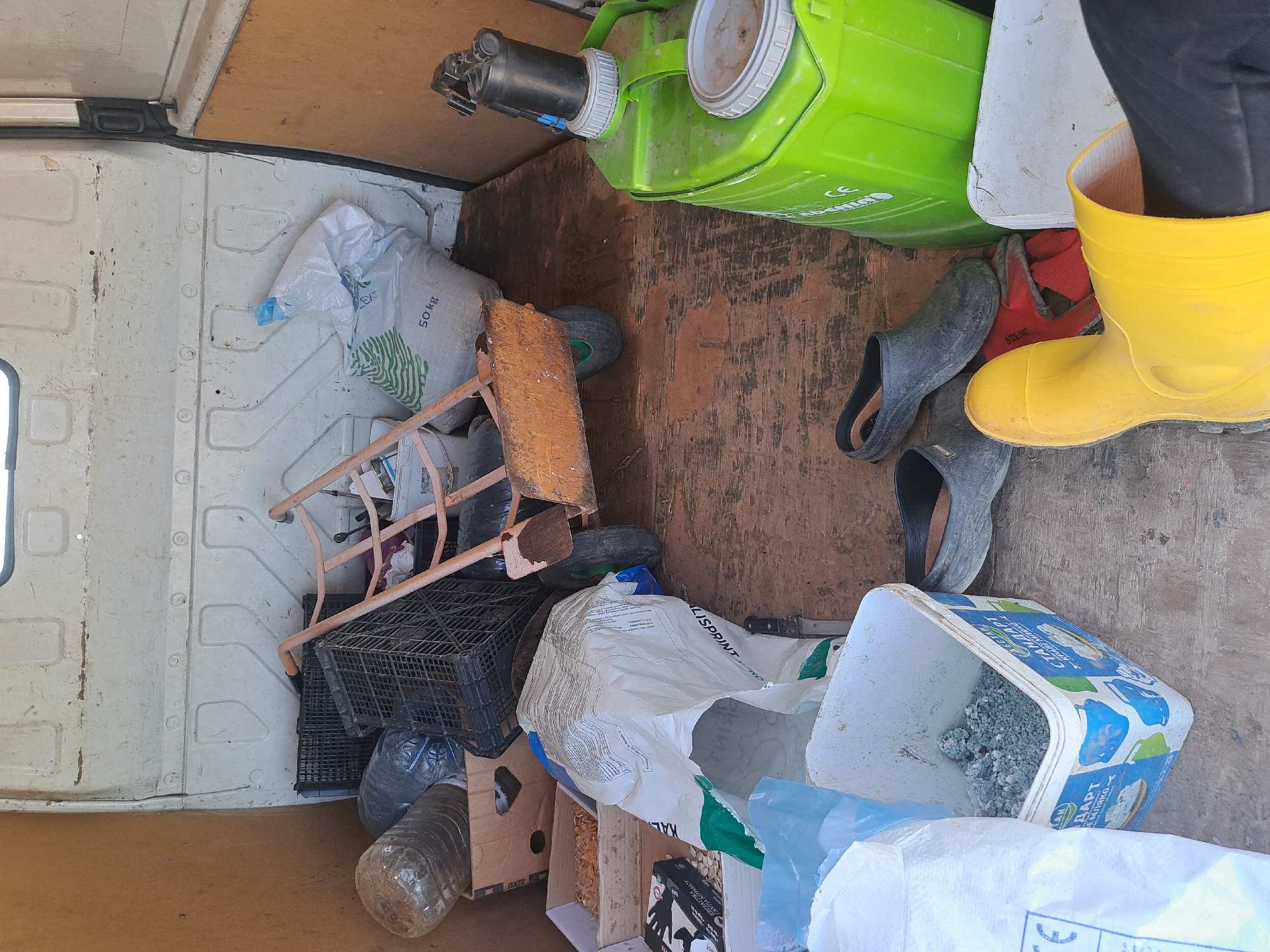 The image depicts the interior of a vehicle, likely a van or truck, in Sofia, Bulgaria, based on the Cyrillic script visible on some of the objects. The cargo area is cluttered with various items, including several large white bags, a green container, a black plastic crate, a ladder, and a box. Two pairs of rubber boots, one yellow and one gray, are visible. A white bucket with blue markings contains gray pellets. The overall impression is one of untidiness and disorganization within the vehicle.