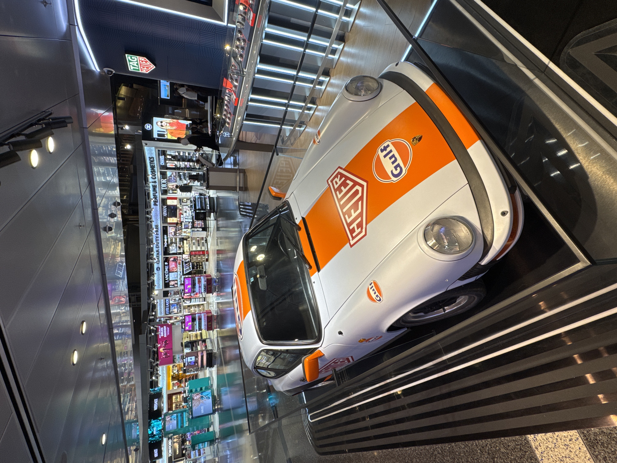 The image depicts a display of a vintage Porsche 911, painted in the iconic Gulf Oil racing livery of white and orange stripes, at the Hamad International Airport in Doha, Qatar. The car is showcased on a platform with reflective black and silver detailing, likely as part of a promotional event. Behind the car, a duty-free shop is visible, featuring shelves stocked with perfumes and cosmetics from various brands such as Dior, Chanel, Lancôme, and Estée Lauder. The Tag Heuer logo is prominently displayed on a blue and white illuminated sign in the background, suggesting a partnership or sponsorship with the watch brand. Several shoppers are milling about in the background, browsing the merchandise. The overall environment suggests a high-end retail space within an international airport.