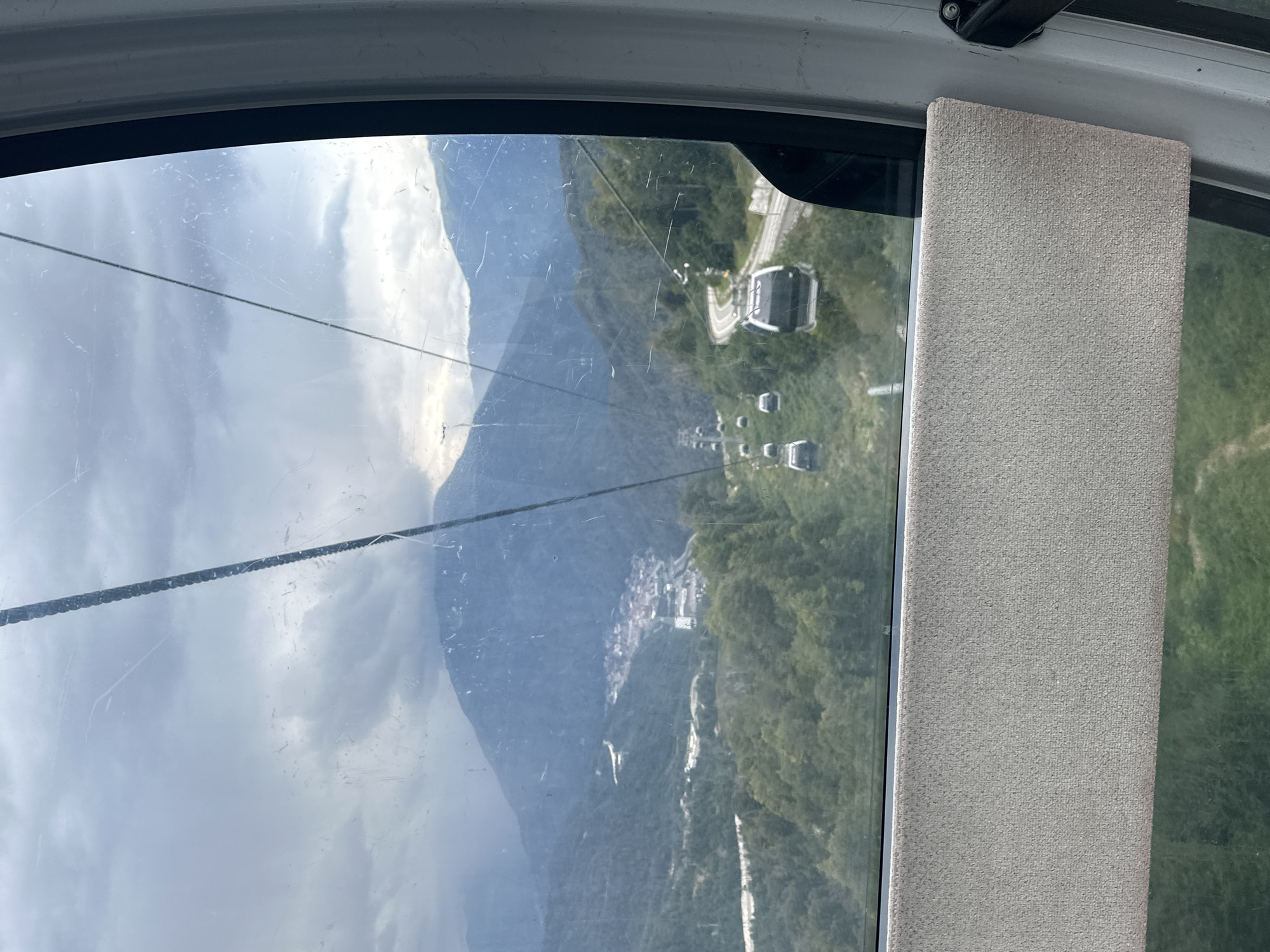 The image captures a view from inside a gondola cabin as it ascends a mountain range. Through the large window, a panoramic landscape unfolds, revealing a valley dotted with buildings and surrounded by lush green slopes. Several other gondola cabins can be seen in the distance, moving along a suspended cable system that snakes its way up the mountain. The sky is overcast with a layer of clouds, creating a somewhat muted light. The foreground of the image shows the interior of the gondola cabin, with a padded armrest visible along the bottom edge. This scene is characteristic of the cable car system in Estosadok, Russia, a popular mountain resort area known for its scenic vistas and recreational activities.