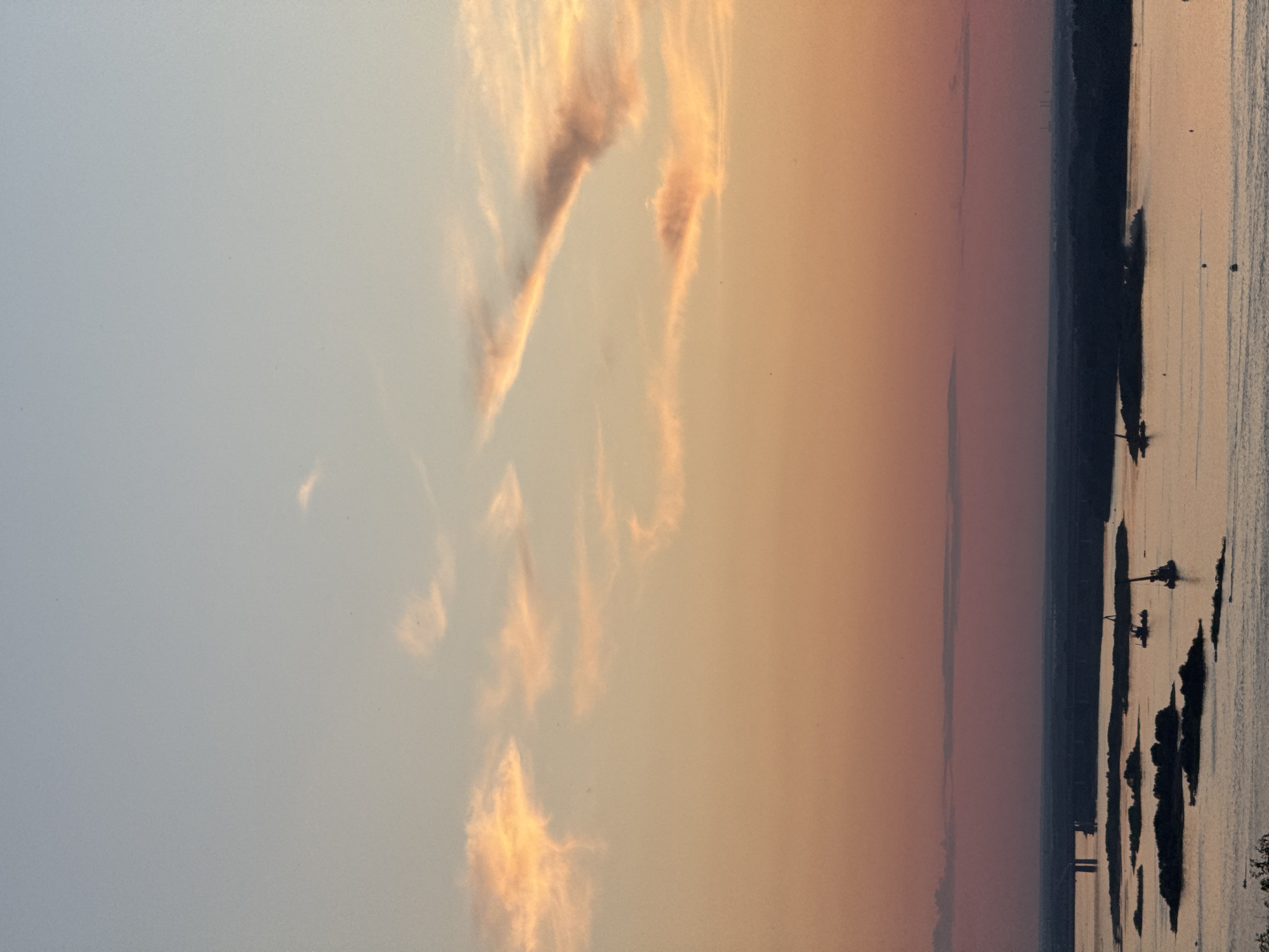 This image captures a twilight scene over the Volga River in Nizhny Novgorod, Russia. The sky displays a gradient of colors, transitioning from a pale blue at the top to shades of orange and pink closer to the horizon, with scattered wispy clouds catching the last light of the sun. In the foreground, the wide expanse of the Volga River reflects the sky's colors, with several dark, silhouetted islands dotting its surface.
On the river, several barges and what appear to be cranes are visible, suggesting ongoing industrial or construction activity. In the distance, a long bridge spans the river, and a dark, tree-covered shoreline forms the far bank. The overall impression is one of a serene, yet industrially active, river landscape at dusk.