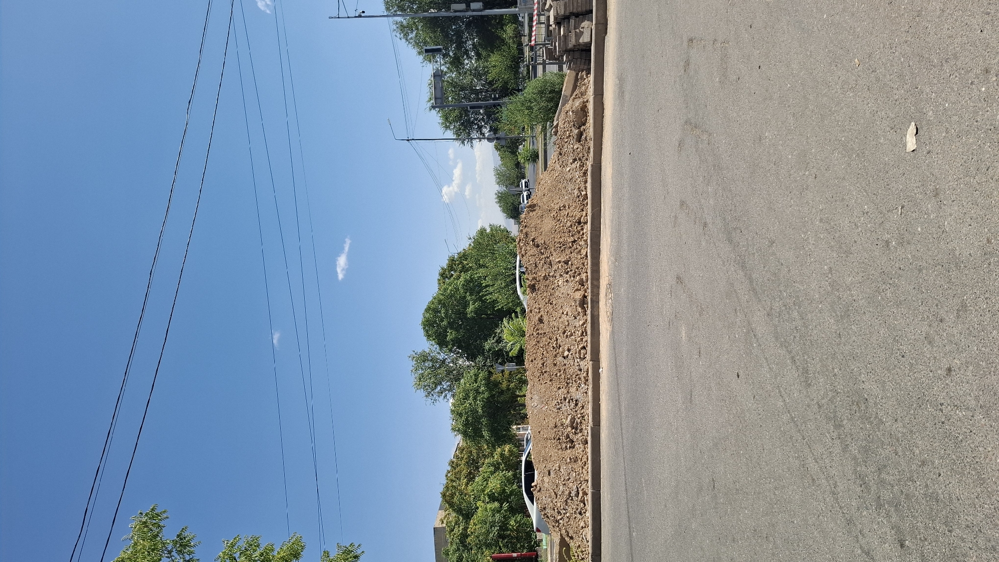 A pile of dirt is located on the side of a road in Shymkent, Kazakhstan. The pile of dirt is in front of a line of trees and a clear blue sky. There are cars on the road and some construction materials are also visible.