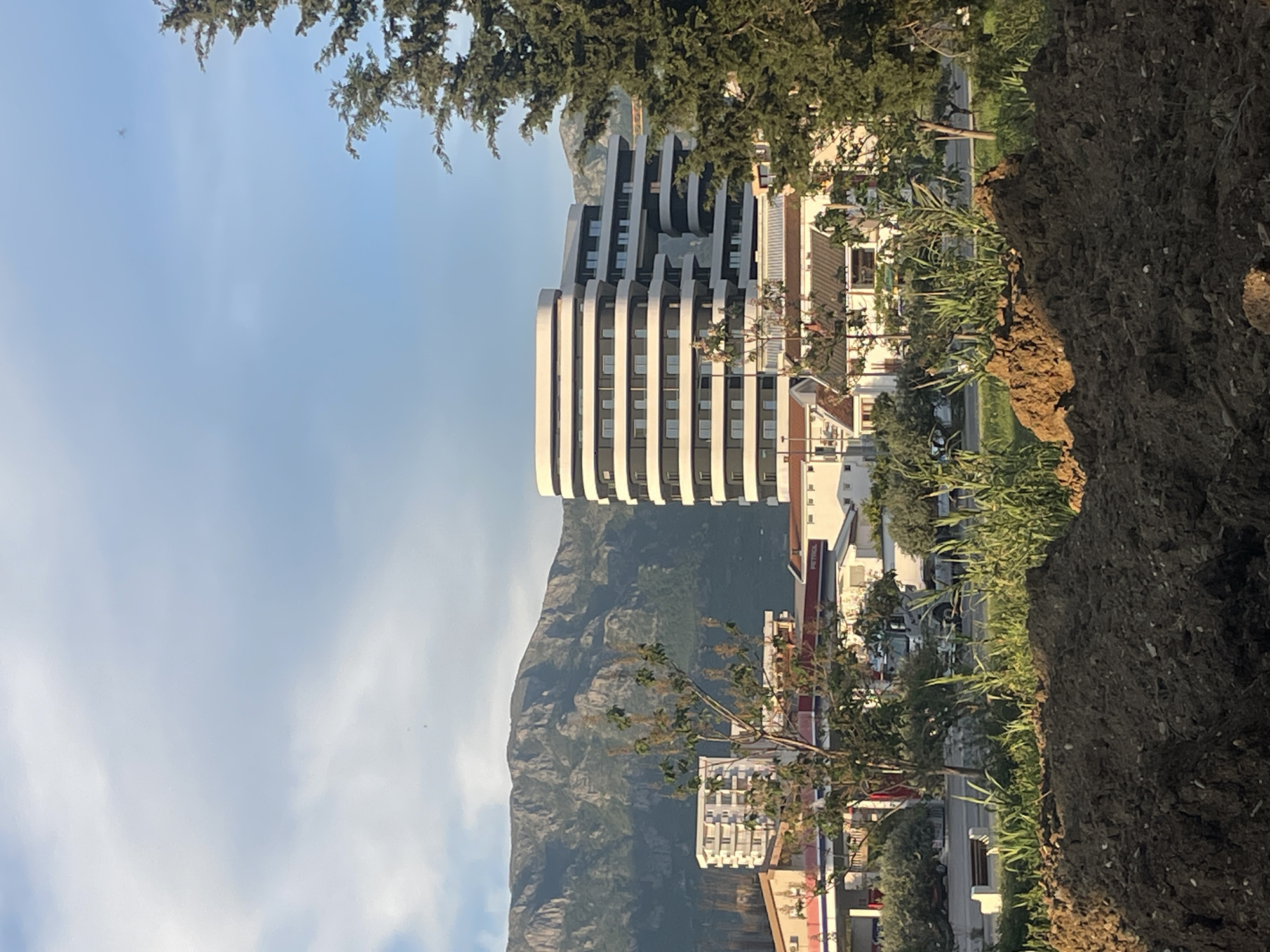 The image presents a landscape view encompassing urban development and natural scenery, set in what appears to be Bar, Montenegro. In the mid-ground, a prominent modern, multi-story building dominates the scene with its distinctive white, curved horizontal facade elements. To the right of this building, other, more traditional structures with terracotta roofs and white walls are visible, nestled among green trees and vegetation.
Behind the urban sprawl, a rugged mountain range stretches across the background, its slopes showing a mix of rocky terrain and verdant growth, all beneath a soft blue sky scattered with delicate white clouds. High in the upper left portion of the sky, a small, distant aircraft can be faintly observed.
In the foreground, towards the bottom right, there is a large mound or bank of dark brown earth, with various green plants and bushes growing both on and behind it. This suggests either a natural embankment or an area of recent construction. Further into the mid-ground on the right, partially obscured by foliage, a building with a visible "PETROL" sign indicates the presence of a gas station. The scene is bathed in the gentle light of day, likely morning or late afternoon, with no harsh shadows. There is no specific activity or event taking place, creating a calm and expansive impression of the location.
