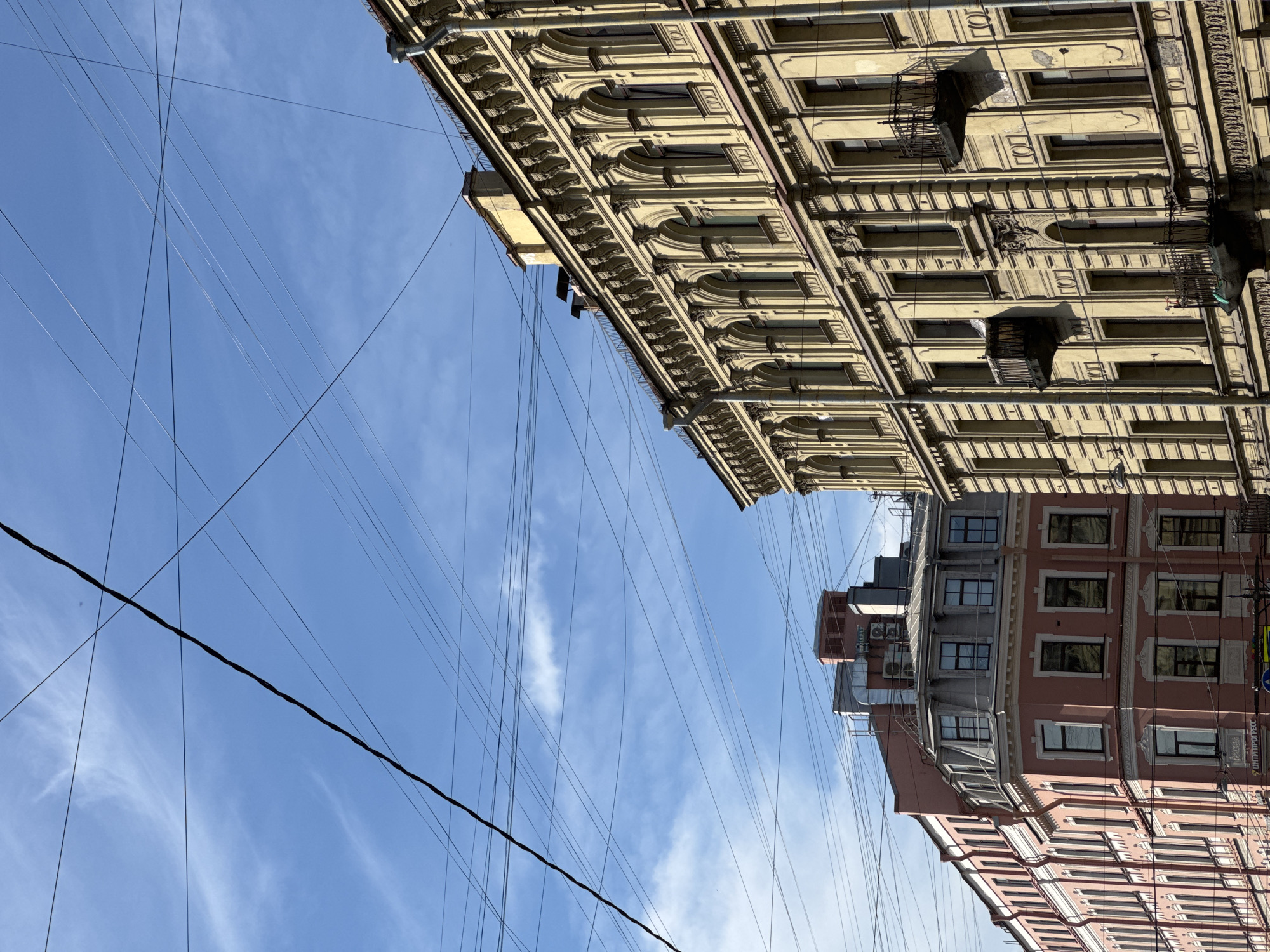 The image depicts an urban scene in Saint Petersburg, Russia, focusing on the architecture of old buildings and a network of overhead wires against a blue sky with thin clouds.
The foreground features the facade of a tall, ornate yellow building with multiple stories. The building exhibits a classical architectural style with a decorative cornice featuring repeating motifs, arched windows, and pilasters. Several small, wrought-iron balconies jut out from the facade.
To the left of the yellow building is another building, this one painted in a salmon pink hue. It is also multi-story and appears to be an older structure with a mansard roof and dormer windows. The pink building has a corner profile, and its facade is adorned with white decorative elements around the windows.
The sky is a bright blue, streaked with wispy white clouds. A complex web of black wires crisscrosses the sky, dominating the upper portion of the image and obscuring parts of the sky and the tops of the buildings. These wires appear to be power or telecommunication lines.
There are no people visible in the image. The scene appears to be a street view looking upwards at the buildings and the sky, suggesting an outdoor, daytime setting. The overall impression is one of historical urban architecture contrasted with the modern utility of overhead wiring.