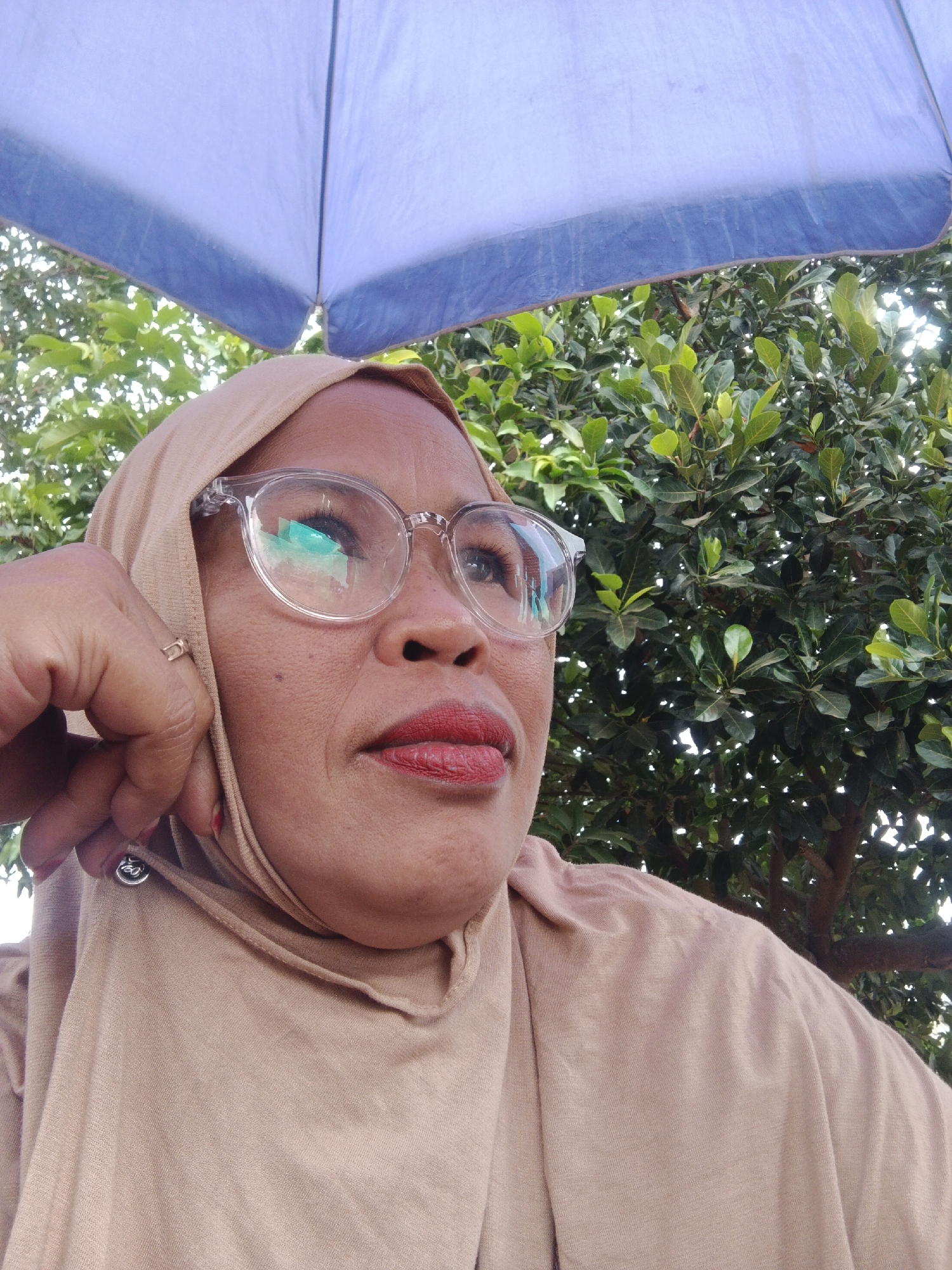 A medium shot captures a woman wearing a tan hijab and clear-rimmed glasses, looking upwards and to the right. Her left hand is raised, with her thumb resting against her cheek and her index finger extended upwards, touching her hijab. She wears a ring on her left ring finger and a circular emblem on her hijab. The woman has red lipstick on and her skin appears to have light makeup. Behind her, a blue umbrella is visible overhead, casting a shadow. Lush green foliage and tree branches fill the background, suggesting an outdoor setting. The lighting indicates daytime, with sunlight filtering through the leaves. This image appears to have been taken outdoors, potentially in a park or garden, possibly within the city of Warungkiara, Indonesia, given the lush vegetation.
