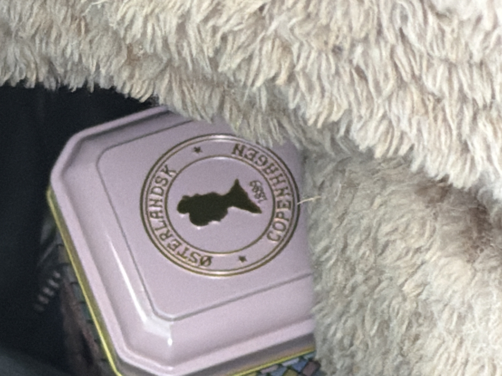 A close-up shot shows the lid of a light pink tin with a gold-trimmed circular emblem. The emblem features the text "ØSTERLANDSK" at the top, a silhouette of a woman's head and shoulders in profile, a small star, "1899" below the silhouette, and "COPENHAGEN" at the bottom. The tin is partially obscured by a fuzzy, light brown blanket. The background is dark and out of focus. The image does not contain enough information to determine its location within Vallensbæk Landsby, Denmark.
