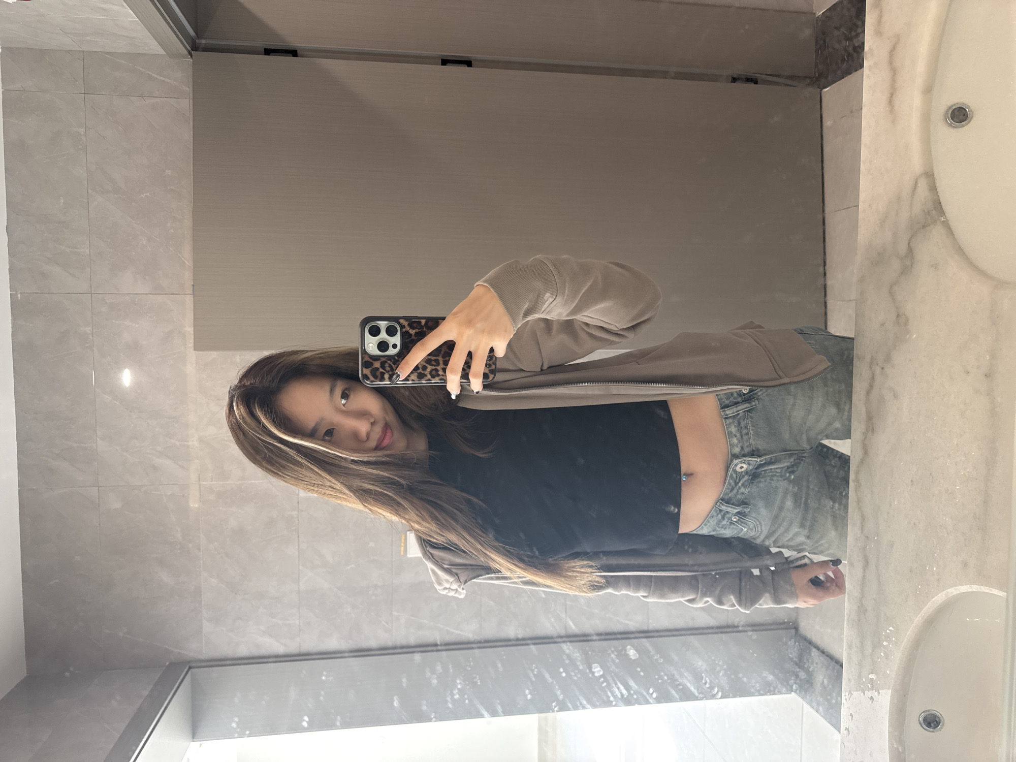 A young woman is taking a selfie in a bathroom. She is wearing a black crop top, light wash jeans, and a beige hoodie. She has long brown hair with blonde highlights. She is holding a phone with a leopard print case. The bathroom has gray tiles, a large mirror, and a white marble countertop with a sink. The location is likely in Nan'an District, China.