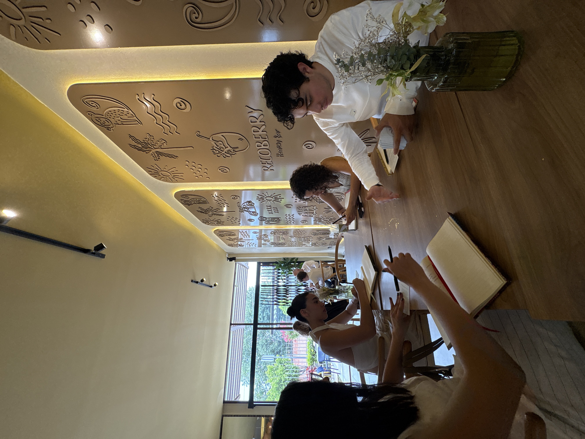 The image shows the interior of Recoberry Health Bar in San Bernardino Tlaxcalancingo, Mexico. Four people are seated at a long wooden table. A man in a white shirt is reaching for a notebook next to a green vase with white flowers. Two women are seated further down the table, appearing to be writing or drawing in notebooks. Another woman is partially visible in the foreground. The ceiling has decorative carvings and recessed lighting. A large window provides a view of a garden.