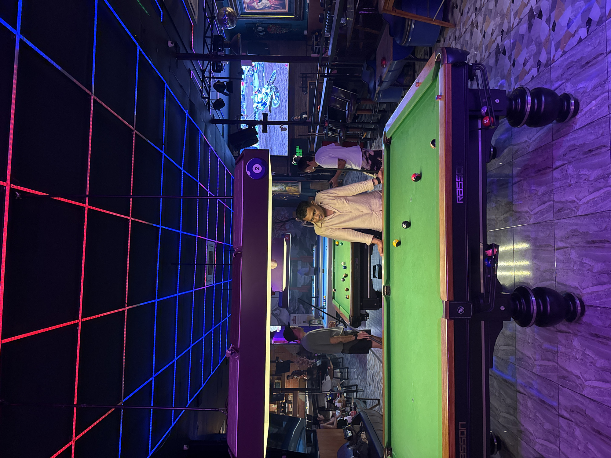 The image depicts a pool hall located in Kuta, Indonesia. The scene is dimly lit, with vibrant red and blue neon lights illuminating the ceiling and casting a colorful glow on the surroundings. In the foreground, a pool table is set up with several balls scattered across its green felt surface. Three men are present in the image. One man, wearing a light-colored long-sleeved shirt, stands by the pool table, appearing to be in the process of playing. Another man, in a gray t-shirt and shorts, holds a pool cue and stands next to the table, observing. A third man, wearing a white t-shirt and patterned shorts, is also near the pool table, seemingly engaged in conversation or observing the game. In the background, another pool table is visible, as well as a bar area with shelves stocked with bottles. A large screen displays a video game, adding to the entertainment atmosphere of the venue. The overall environment suggests a casual and lively setting, typical of a bar or entertainment establishment.